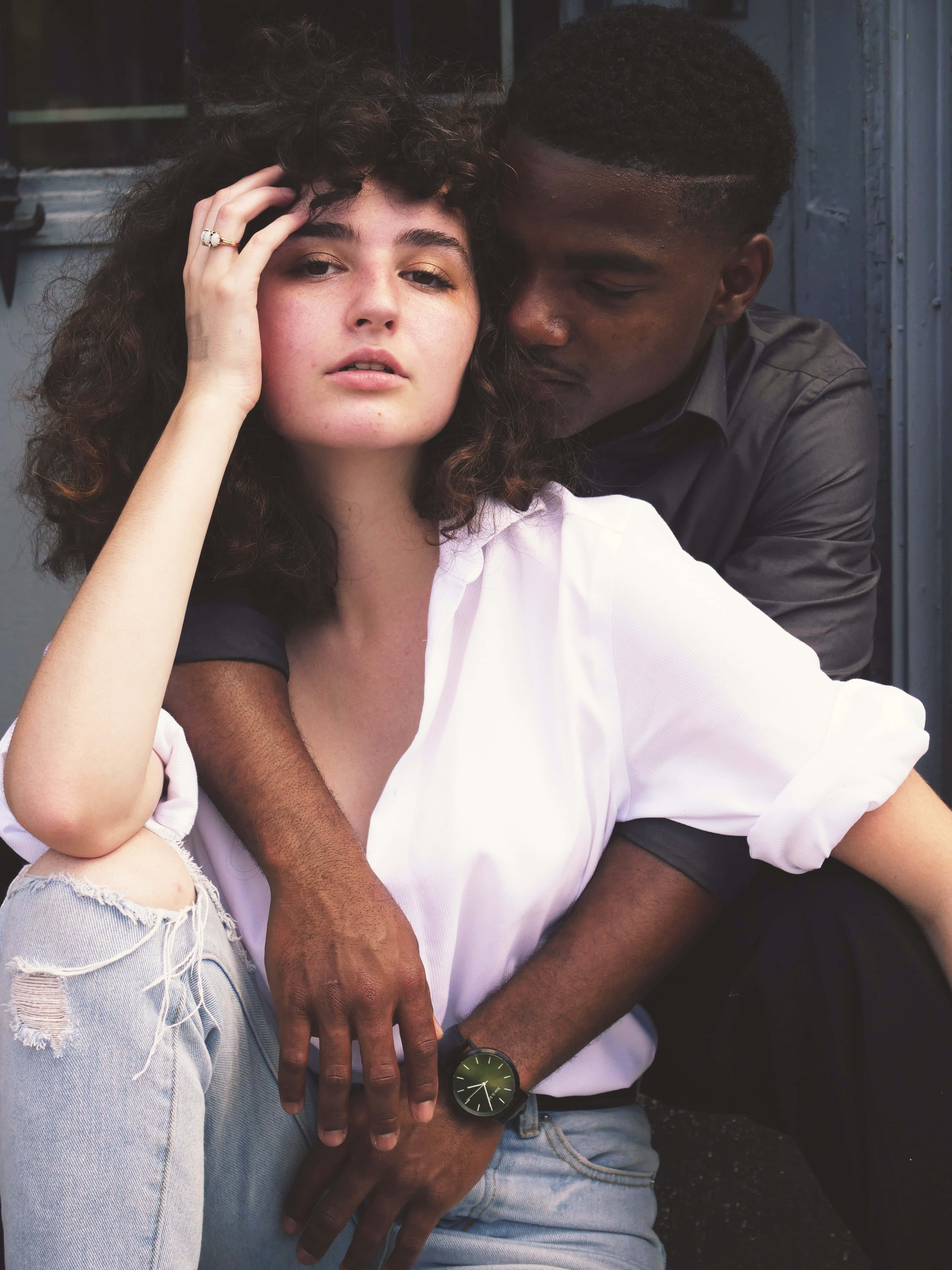 Interracial 桌布 壁紙 — 一位年輕的捲髮女子若有所思地坐著，一名男子從背後擁抱她，構成一幅自然的城市肖像。