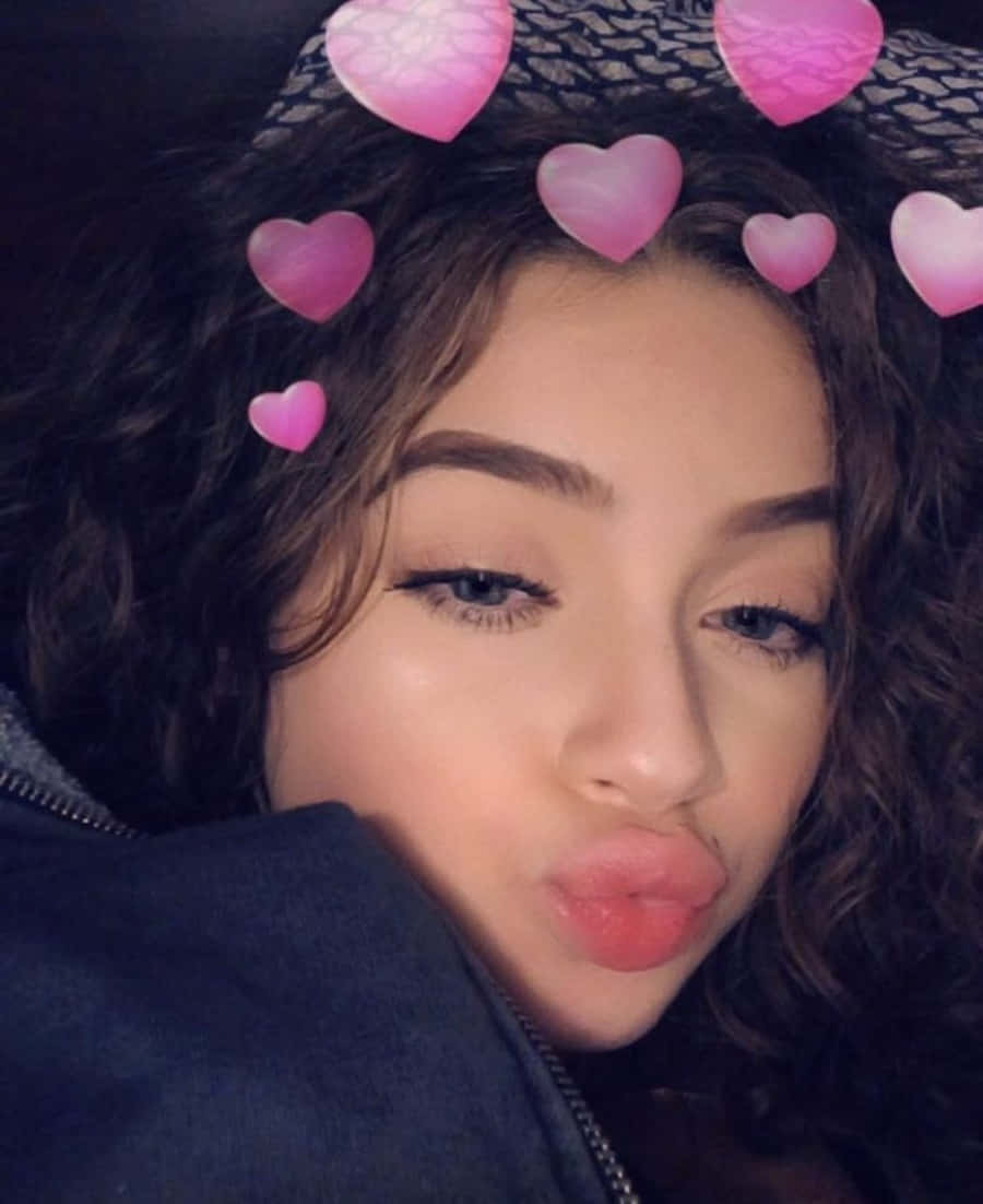 Snapchat 桌布 壁紙 — 一位年輕的捲髮女性在自拍中，被漂浮的粉紅色心形包圍，飛吻。