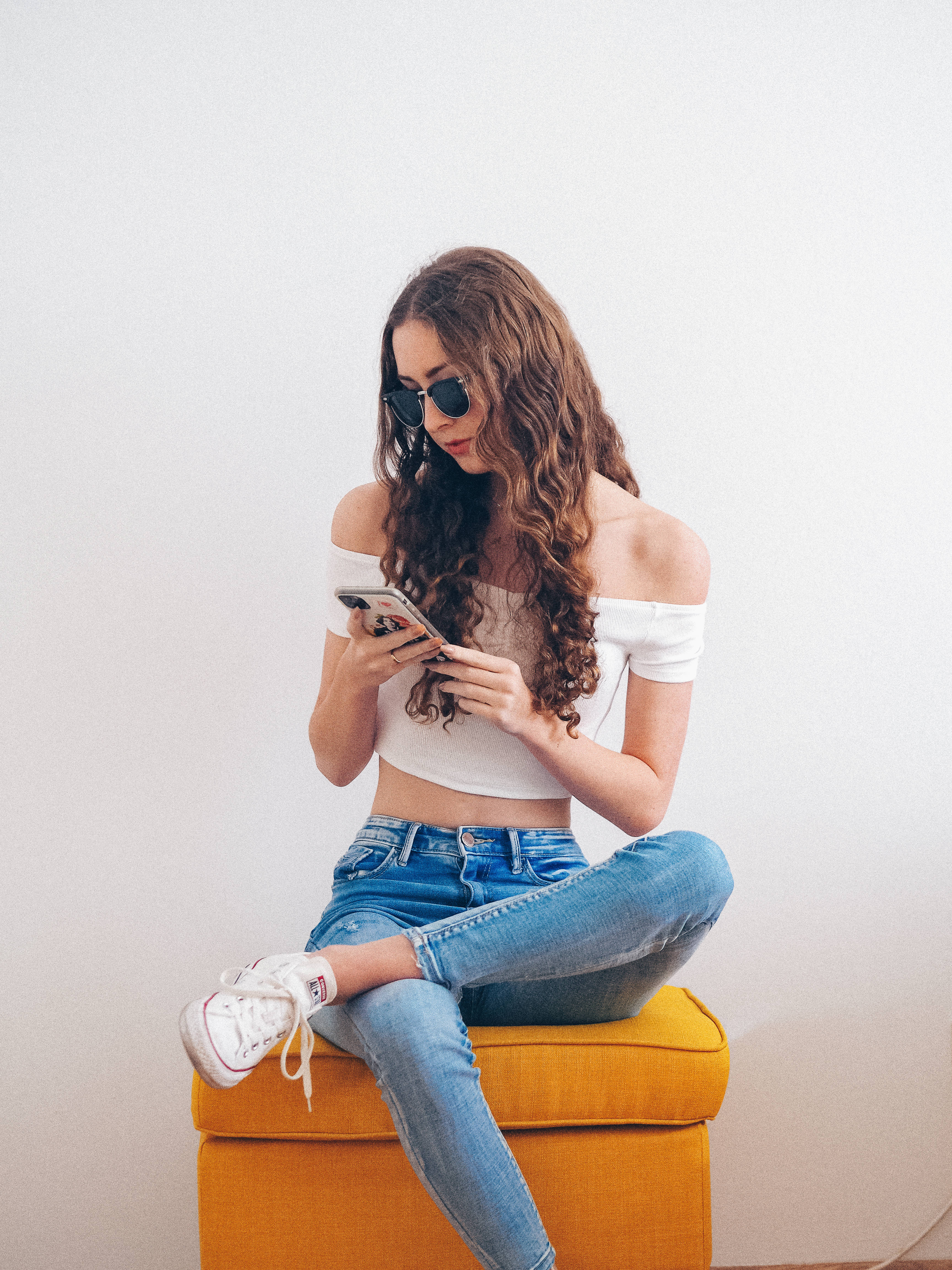 Trendy 桌布 壁紙 — 一位年輕的捲髮女性戴著太陽眼鏡，盤腿坐在鮮黃色的軟凳上，專注於手機。