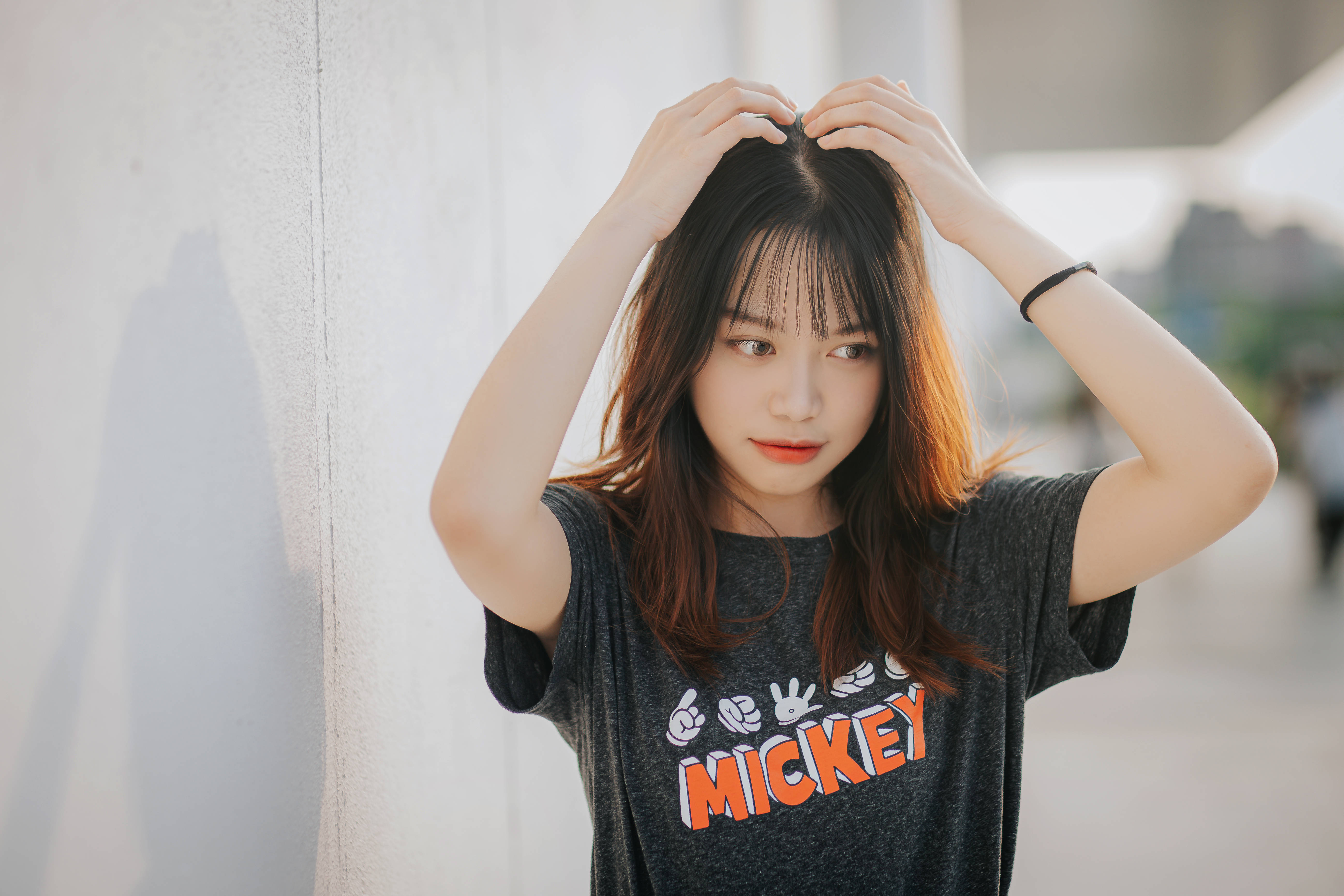 女孩 桌布 壁紙 — 一位年輕的棕髮女性穿著米奇老鼠T恤，輕輕地將頭髮觸碰在明亮的白色牆壁上。