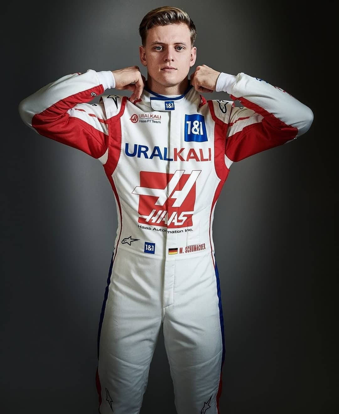 mick schumacher 桌布 壁紙 — 一位年輕的男性賽車手穿著白色、紅色和藍色的Haas F1車隊賽車服，在黑暗的攝影棚背景下自信地擺姿勢。