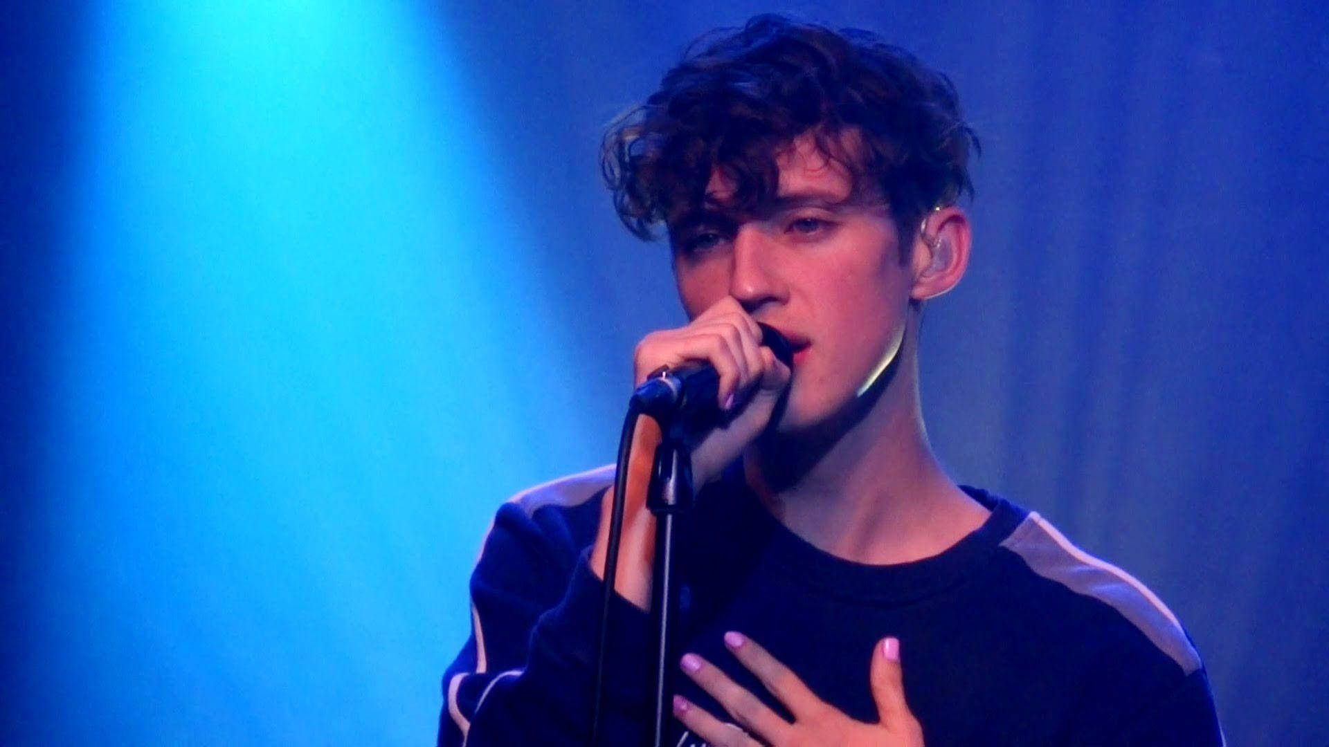 Sivan 桌布 壁紙 — 一位年輕的男歌手，頭髮捲曲，在充滿活力的藍色舞台燈光下熱情地對著麥克風演唱。