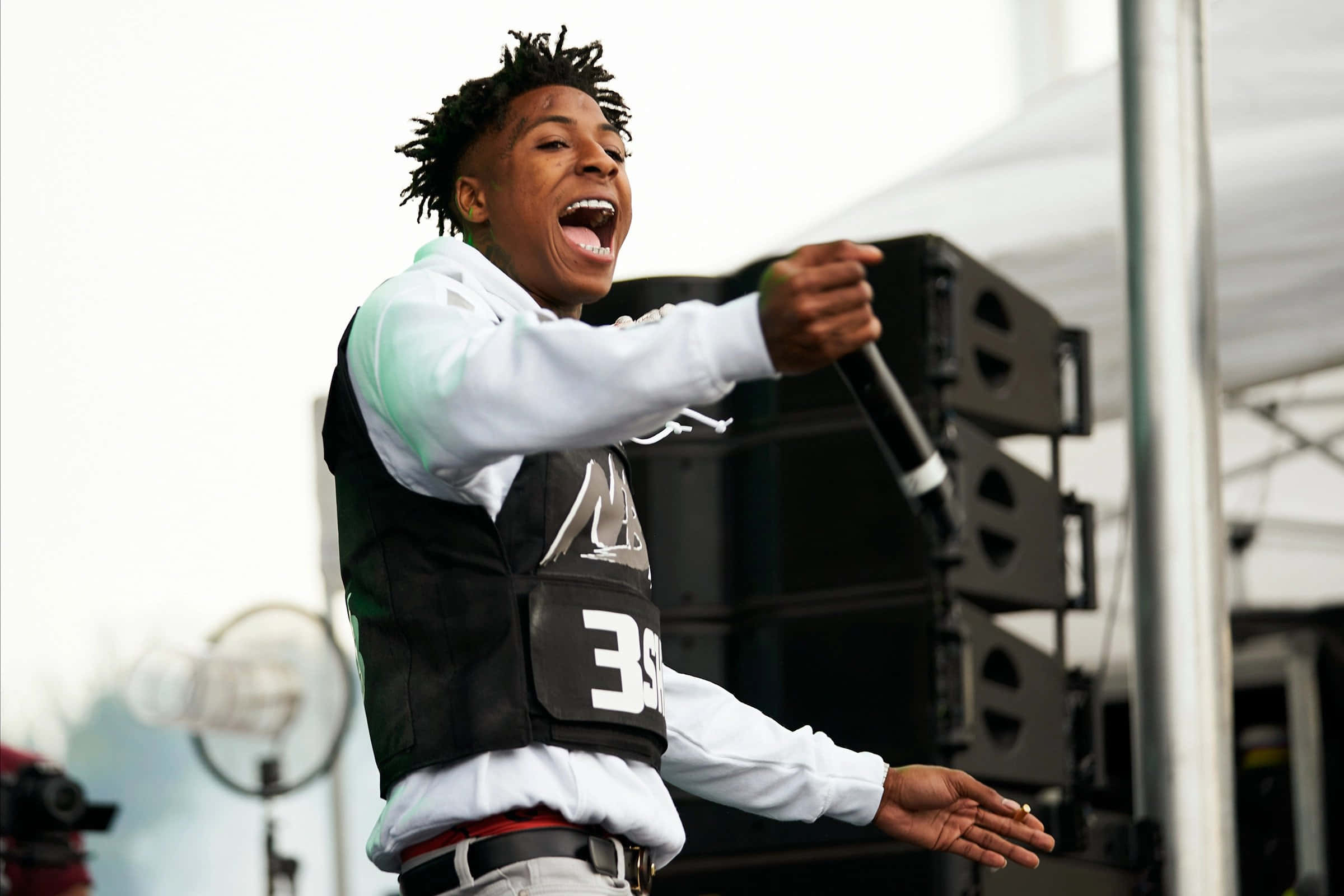 NBA YoungBoy 桌布 壁紙 — 一位年輕的表演者留著辮子，在白色帳篷下的舞台上熱情地對著麥克風唱歌。