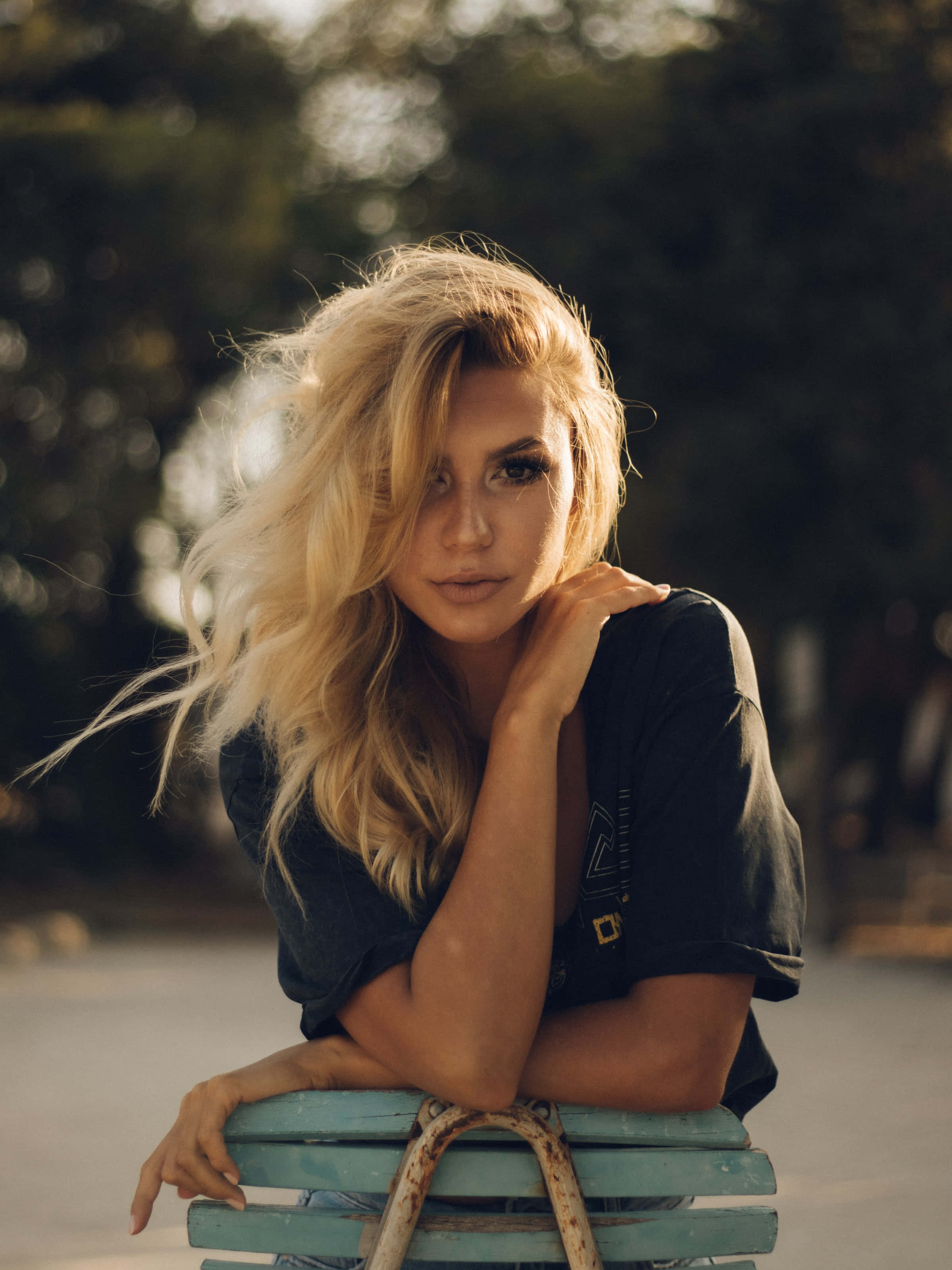 Blonde Woman 桌布 壁紙 — 一位年輕的金髮女子，頭髮被風吹亂，坐在陽光明媚的公園裡一張風化的藍色長椅上，手托著下巴。