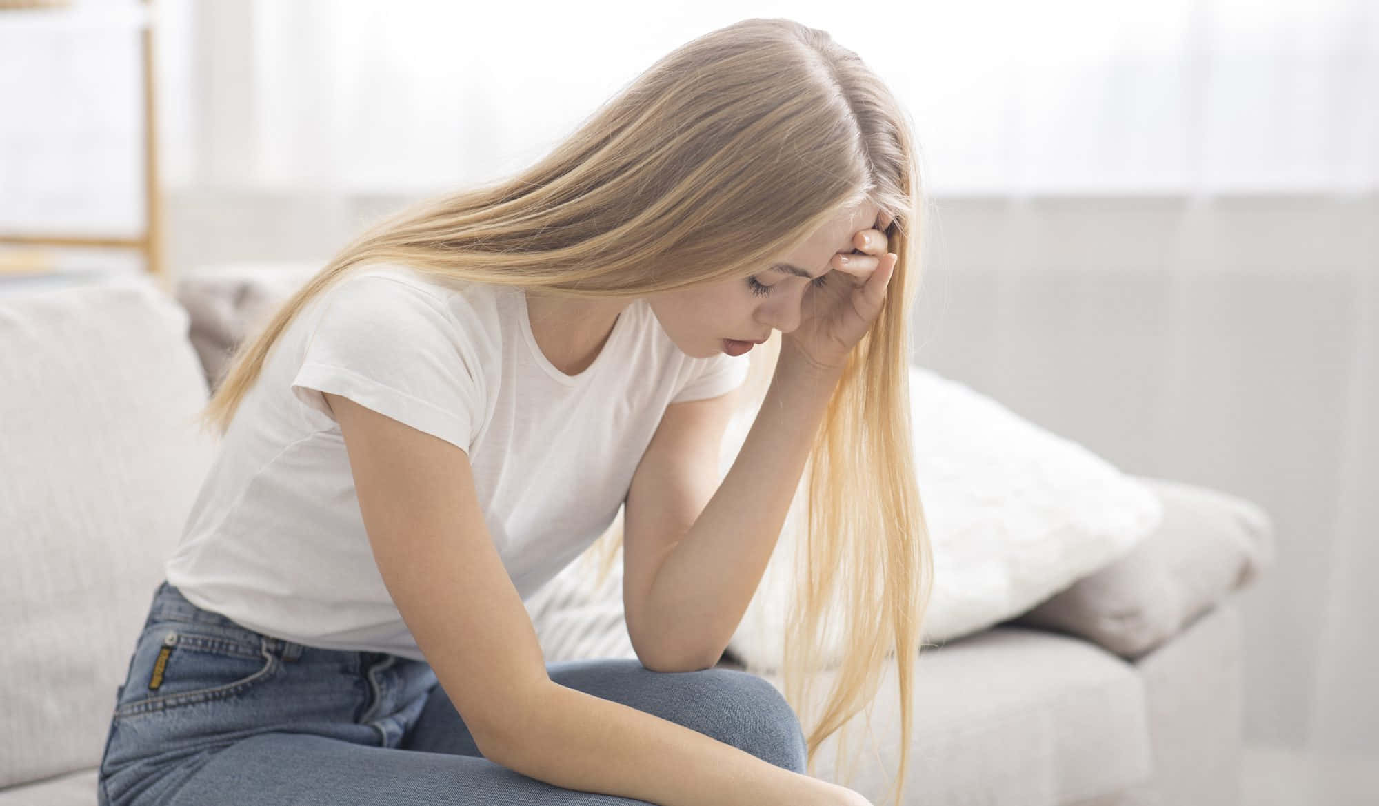 Teen Girl 桌布 壁紙 — 一位年輕的長金髮女性癱坐在沙發上，痛苦地抱著頭。