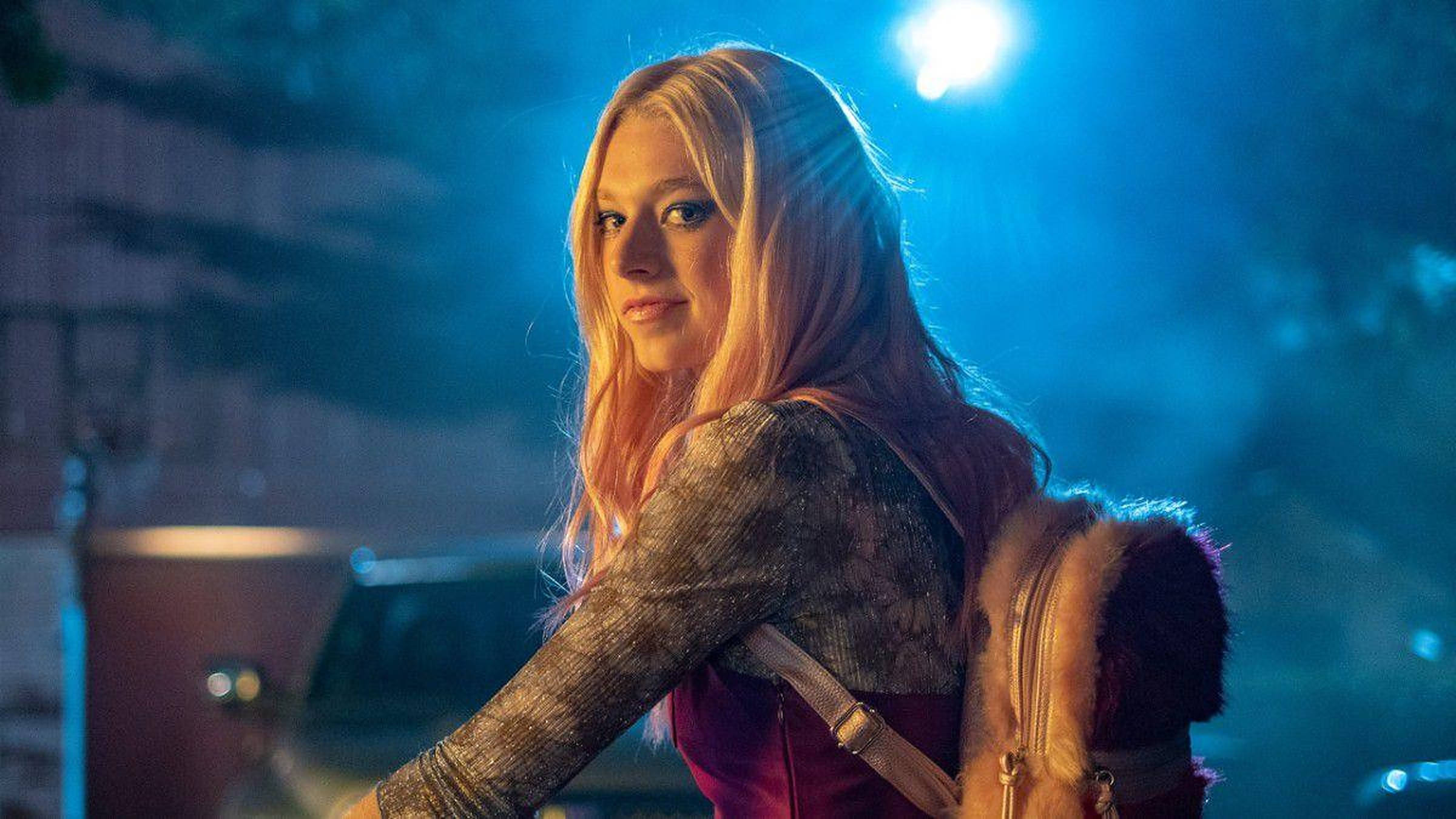 Hunter Schafer 桌布 壁紙 — 一位年輕的長髮金髮女性在夜晚站在明亮的藍色聚光燈下，穿著質感上衣和毛邊背包。
