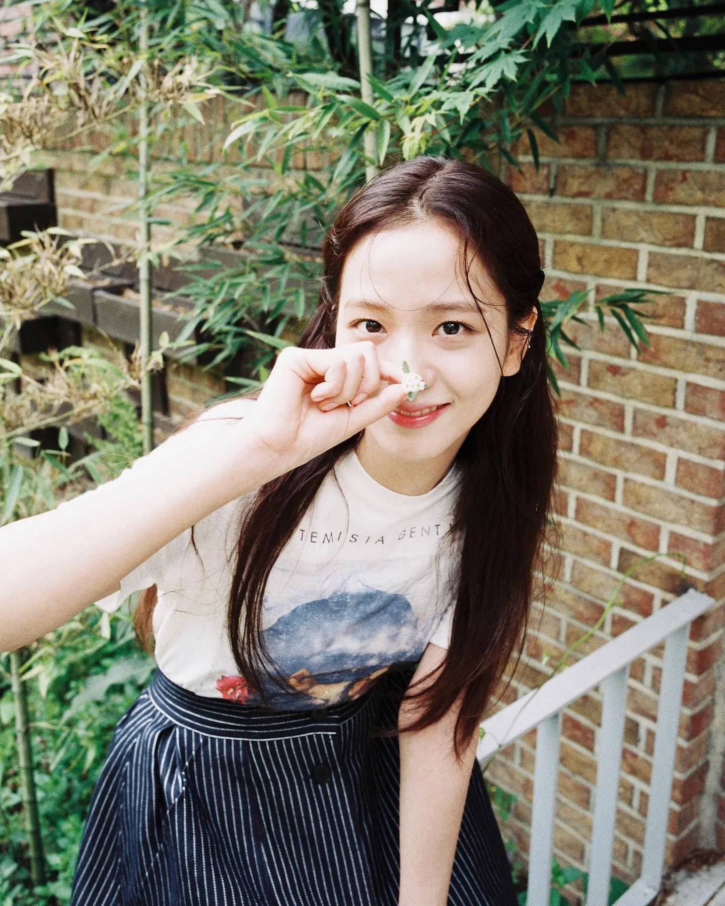 jisoo 可愛 桌布 壁紙 — 一位年輕的長黑髮女性，在磚牆和竹子的背景下，將一朵小白花靠近鼻子，溫柔地微笑。