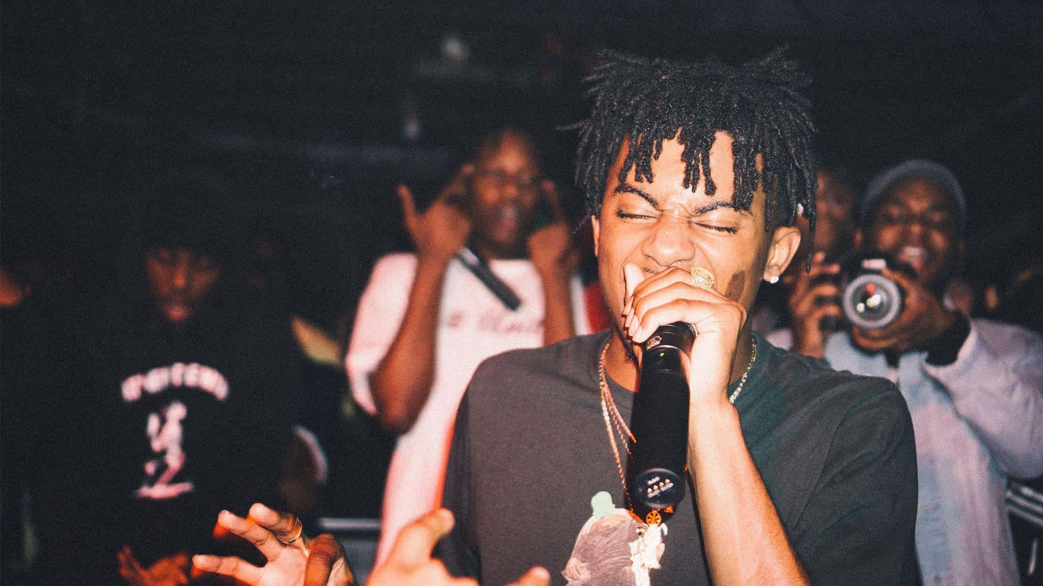 Playboi Carti 桌布 壁紙 — 一位年輕的雷鬼辮說唱歌手在昏暗的俱樂部燈光下對著麥克風熱情表演，身後是投入的觀眾。
