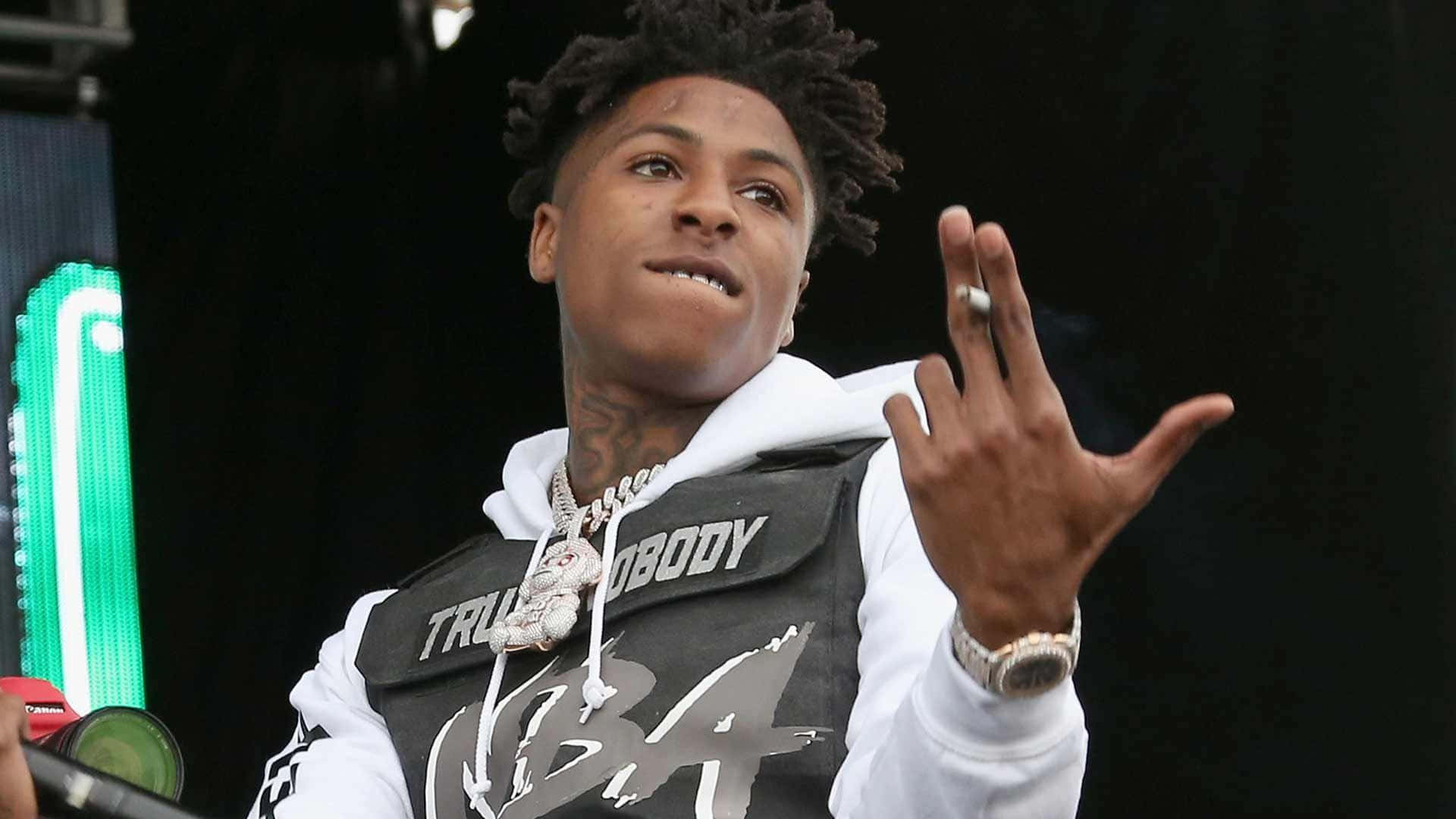 NBA YoungBoy 桌布 壁紙 — 一位年輕的雷鬼辮饒舌歌手在舞台上自信地做手勢，身穿白色連帽衫和帶有粗體字樣的黑色背心。
