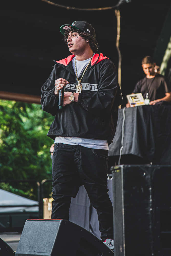 Shoreline Mafia 桌布 壁紙 — 一位年輕的饒舌歌手穿著黑色外套和帽子，在舞台上拿著麥克風表演，身後有綠色樹葉。