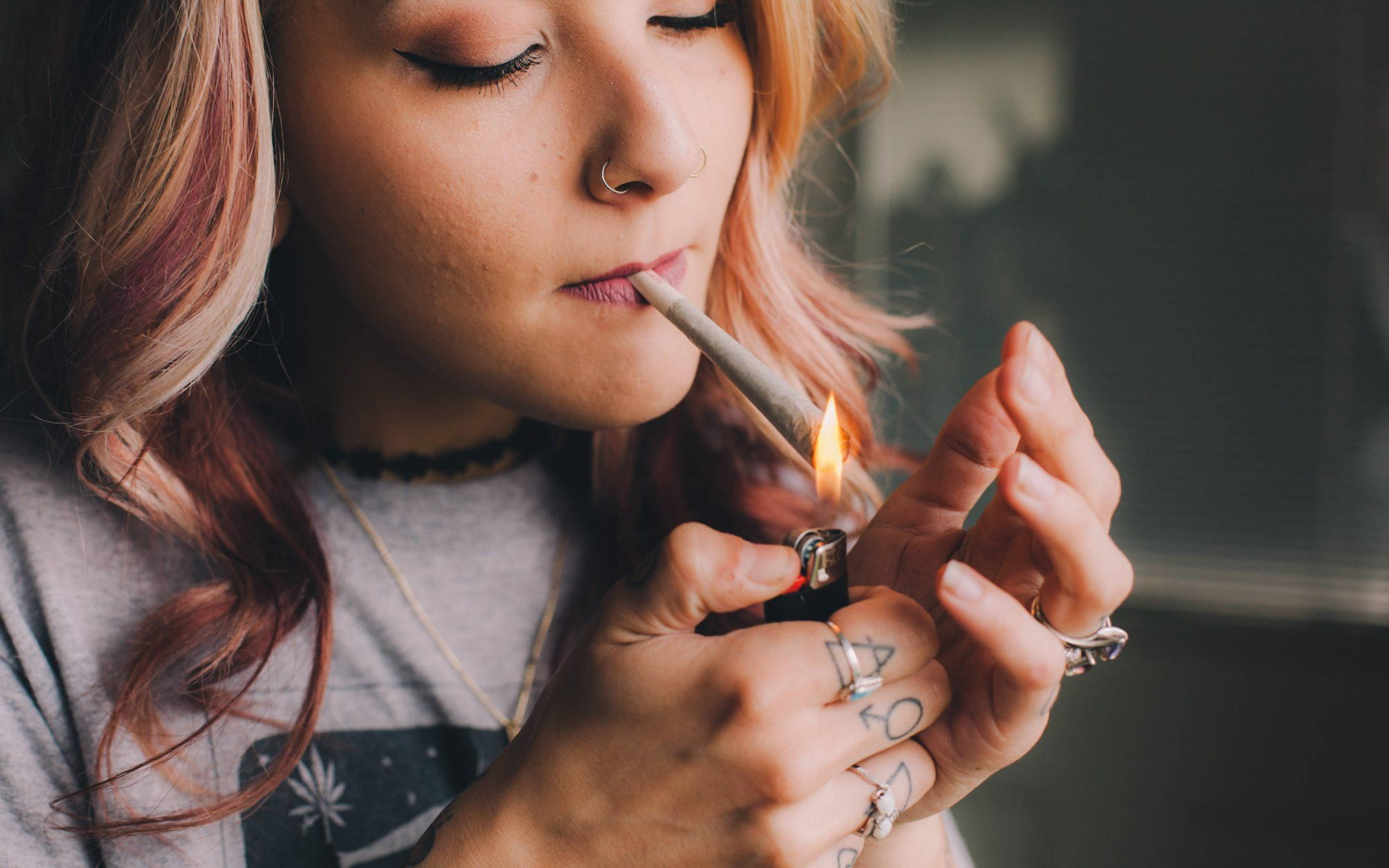吸大麻 桌布 壁紙 — 一位年輕粉紅髮女性在昏暗的室內環境中用打火機點燃一支大麻煙。