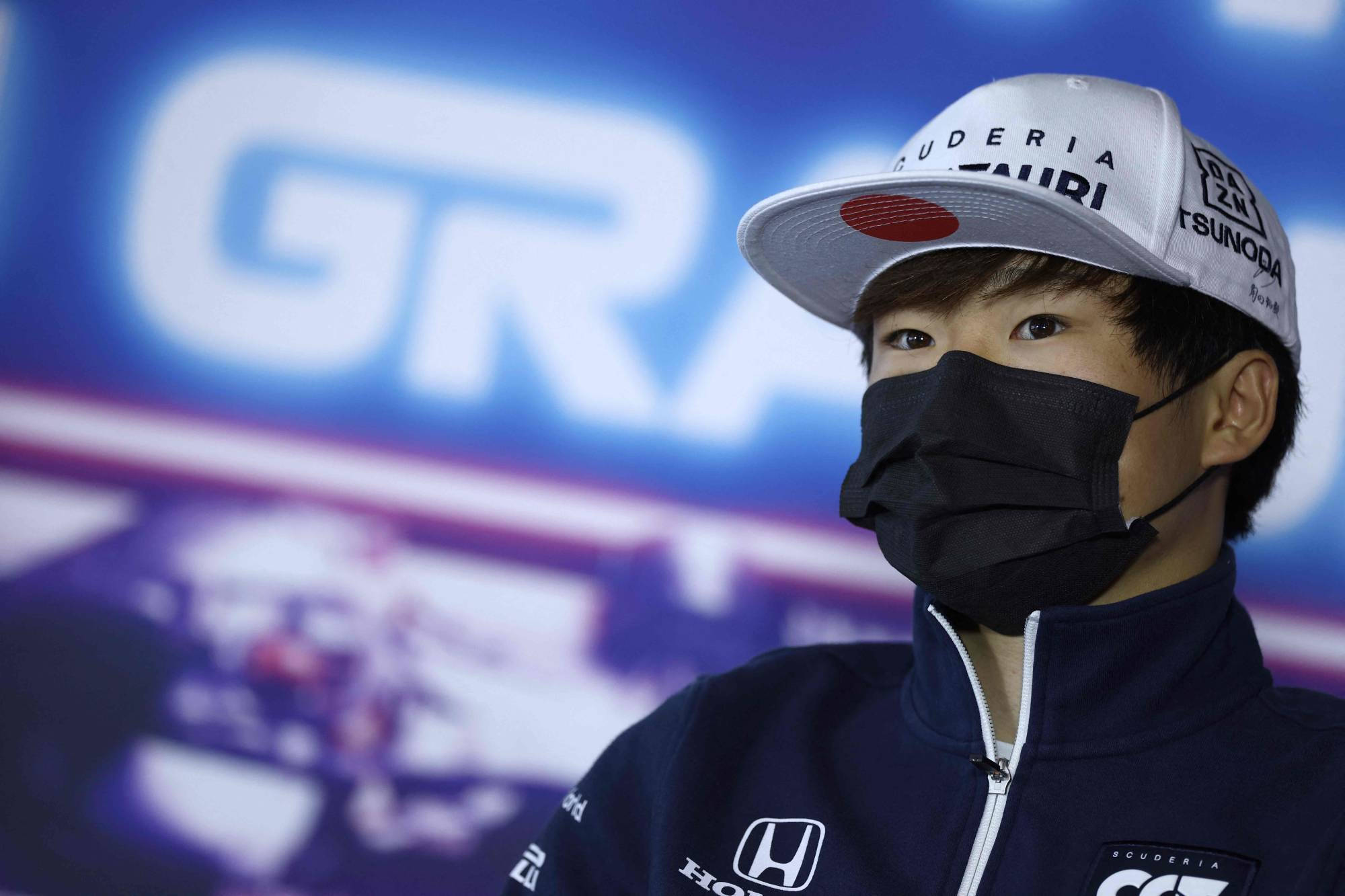 Honda 桌布 壁紙 — 一位年輕賽車手戴著 Scuderia AlphaTauri 帽子和黑色口罩，在模糊的藍白品牌背景前若有所思地凝視。
