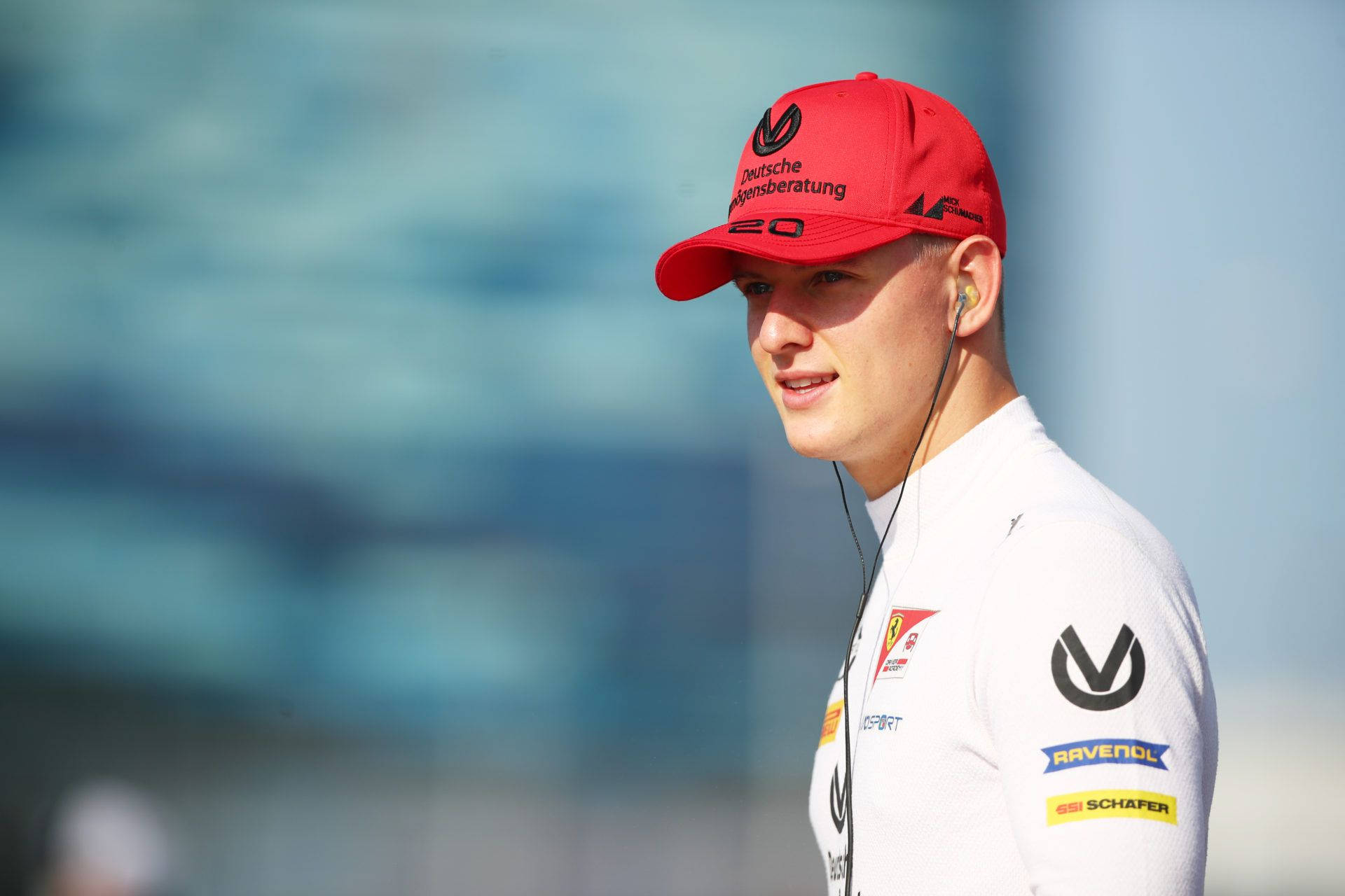mick schumacher 桌布 壁紙 — 一位年輕賽車手戴著紅色帽子，身穿白色隊服外套，在明亮的日光下神情專注地站立。