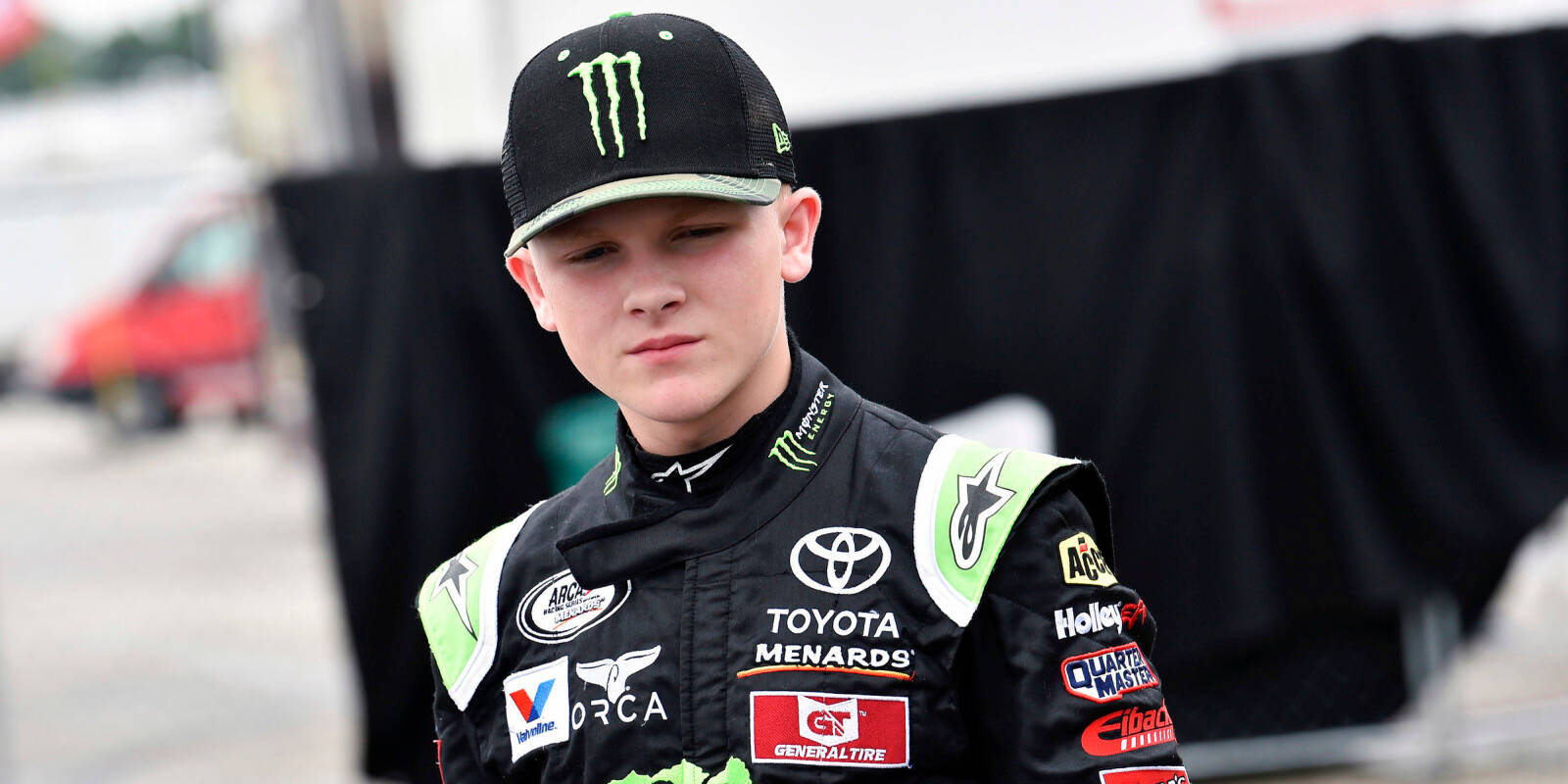 Ty Gibbs 桌布 壁紙 — 一位年輕賽車手穿著黑色和綠色的 Toyota Menards 賽車服和帽子，神情專注地站立。