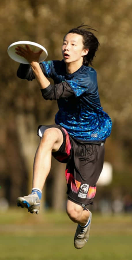 Athlete 桌布 壁紙 — 一位年輕運動員在陽光明媚的公園中躍起，在模糊的樹木背景下，專注地接住一個白色飛盤。