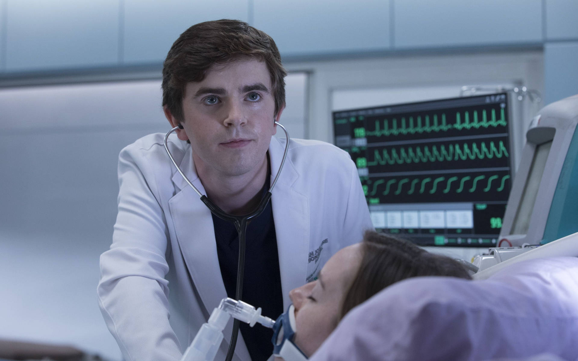 Freddie Highmore 桌布 壁紙 — 一位年輕醫生俯身靠近病床上的病人，脖子上掛著聽診器，身後有顯示綠色心跳線的監視器。