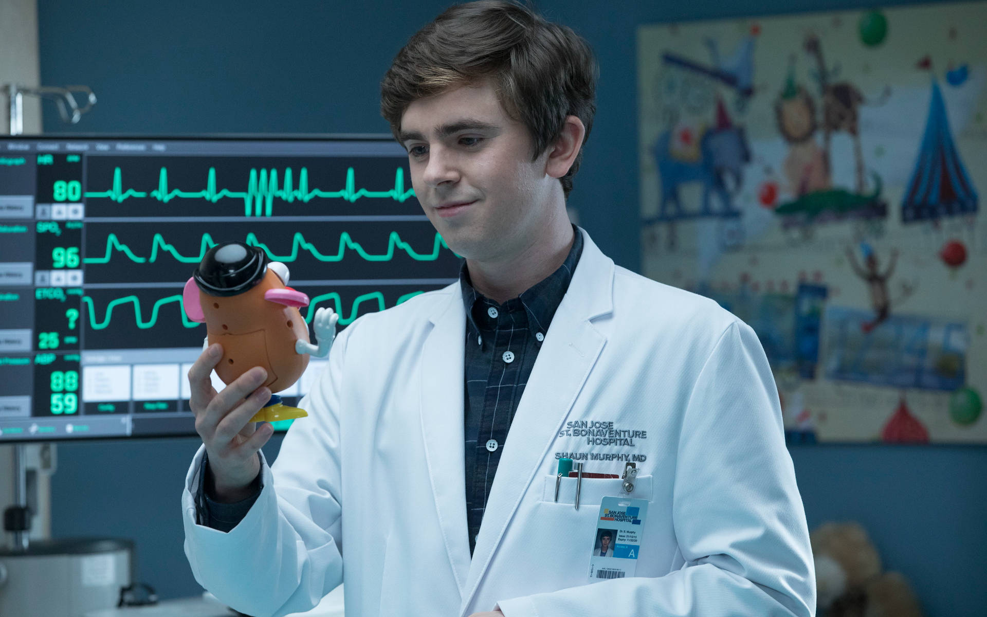 Freddie Highmore 桌布 壁紙 — 一位年輕醫生穿著白袍，在有心臟監測儀的病房裡拿著一個玩具機器人。