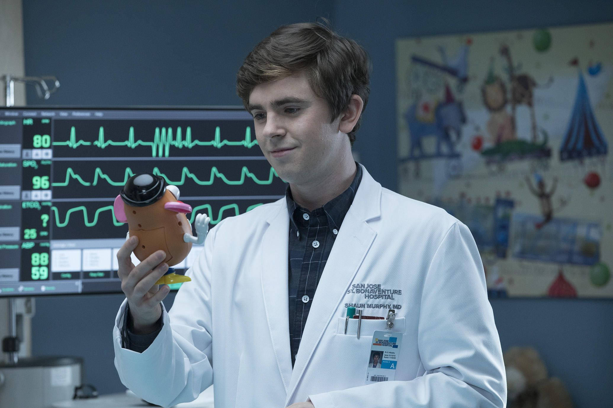 The Good Doctor 桌布 壁紙 — 一位年輕醫生穿著白袍，在病房裡拿著玩具聽診器，身後有心臟監護儀。