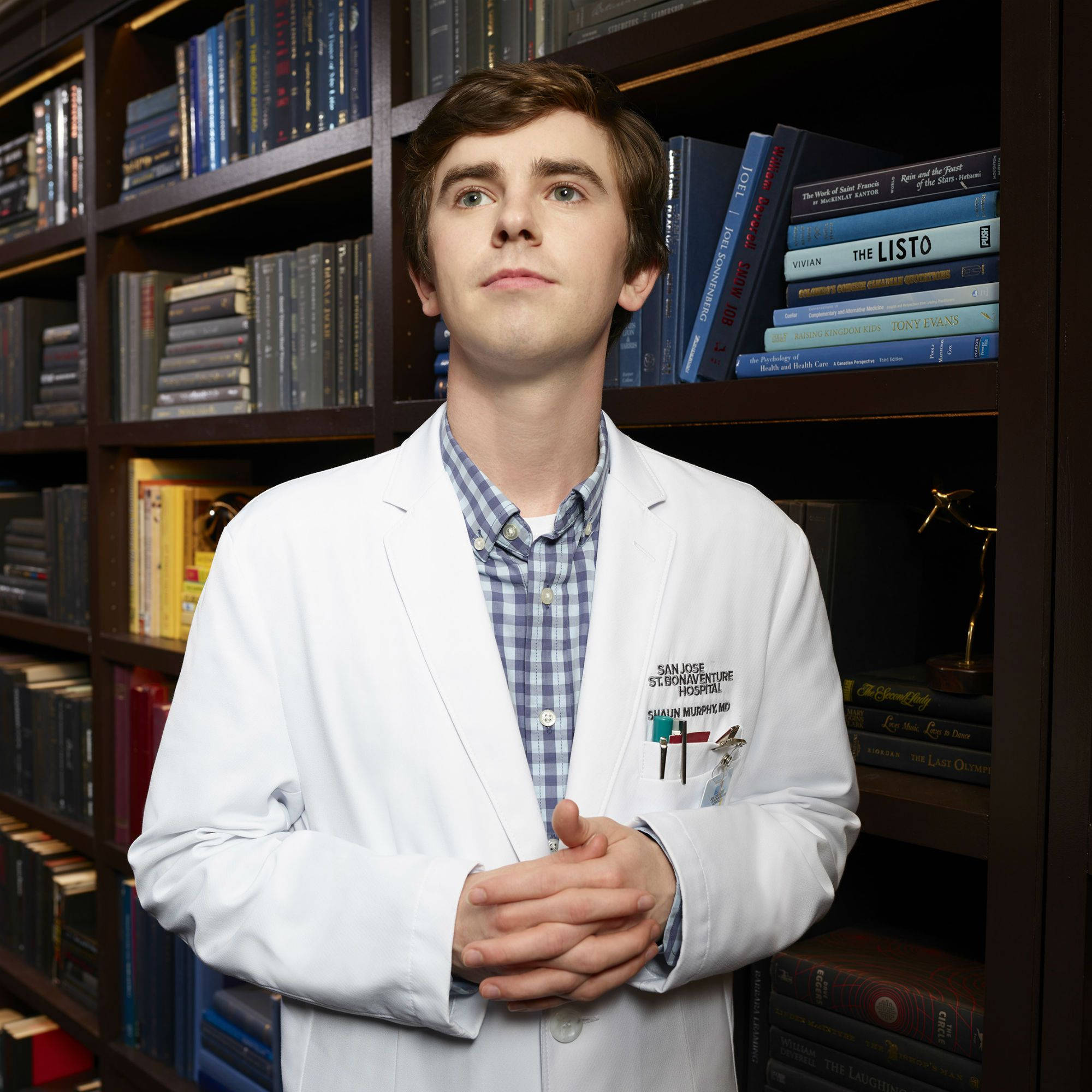 Freddie Highmore 桌布 壁紙 — 一位年輕醫生身穿白色實驗室外套，自信地站在裝滿醫學文獻和獎項的書架前。