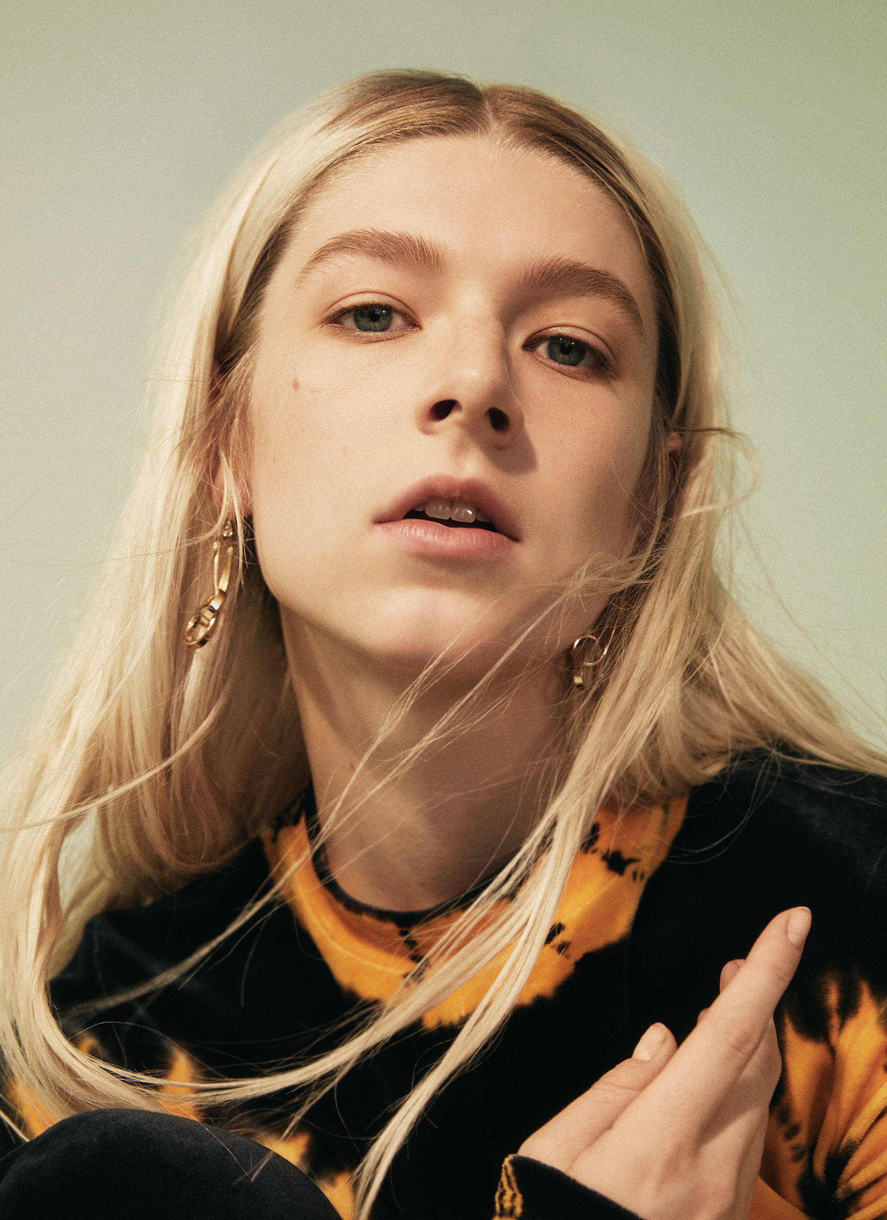 Hunter Schafer 桌布 壁紙 — 一位年輕金髮女子戴著金色圓環耳環，直視鏡頭，身穿黑色和橙色紮染上衣。