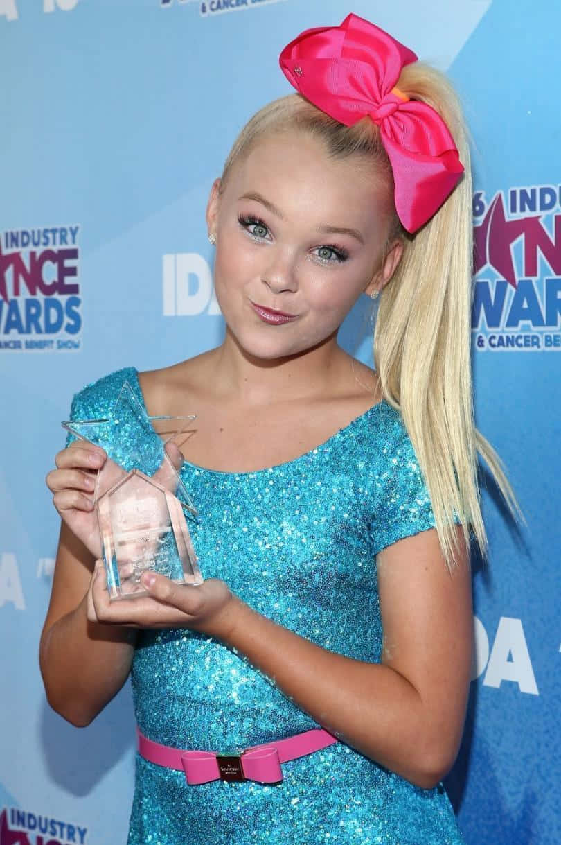 JoJo Siwa 桌布 壁紙 — 一位年輕金髮女孩戴著巨大粉紅色蝴蝶結，在藍色活動背景前拿著透明獎盃。