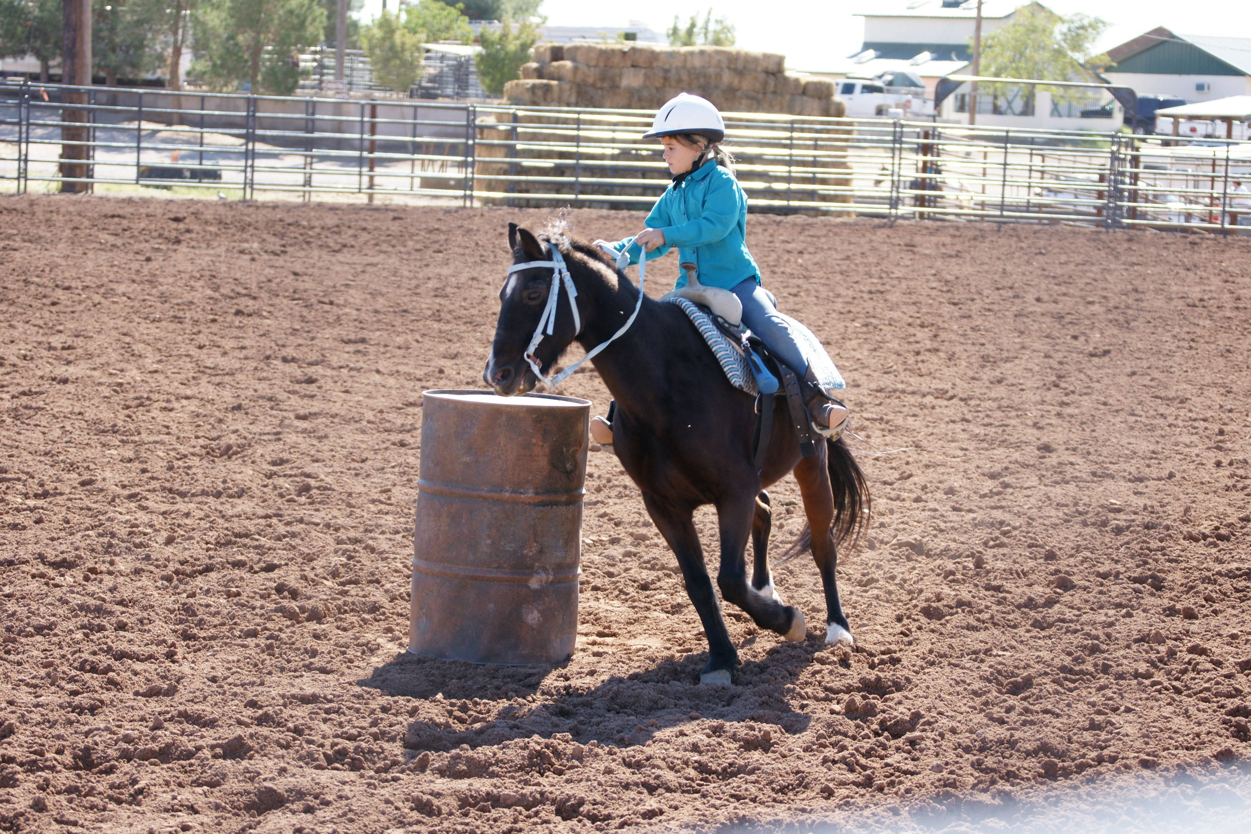 Barrel Racing 桌布 壁紙 — 一位年輕騎士身穿青綠色夾克和白色頭盔，在陽光明媚的泥地競技場中引導一匹深色馬繞著生鏽的桶子轉圈。