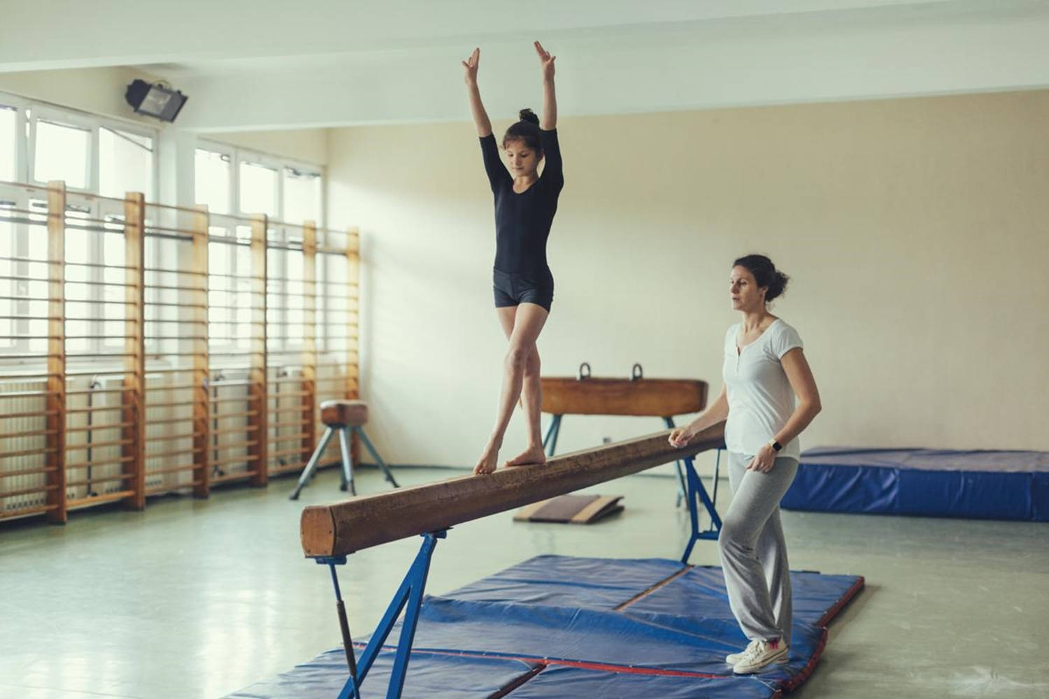 Stretching 桌布 壁紙 — 一位年輕體操運動員在明亮的體育館中優雅地在木製平衡木上保持平衡，而教練則專注地觀看。