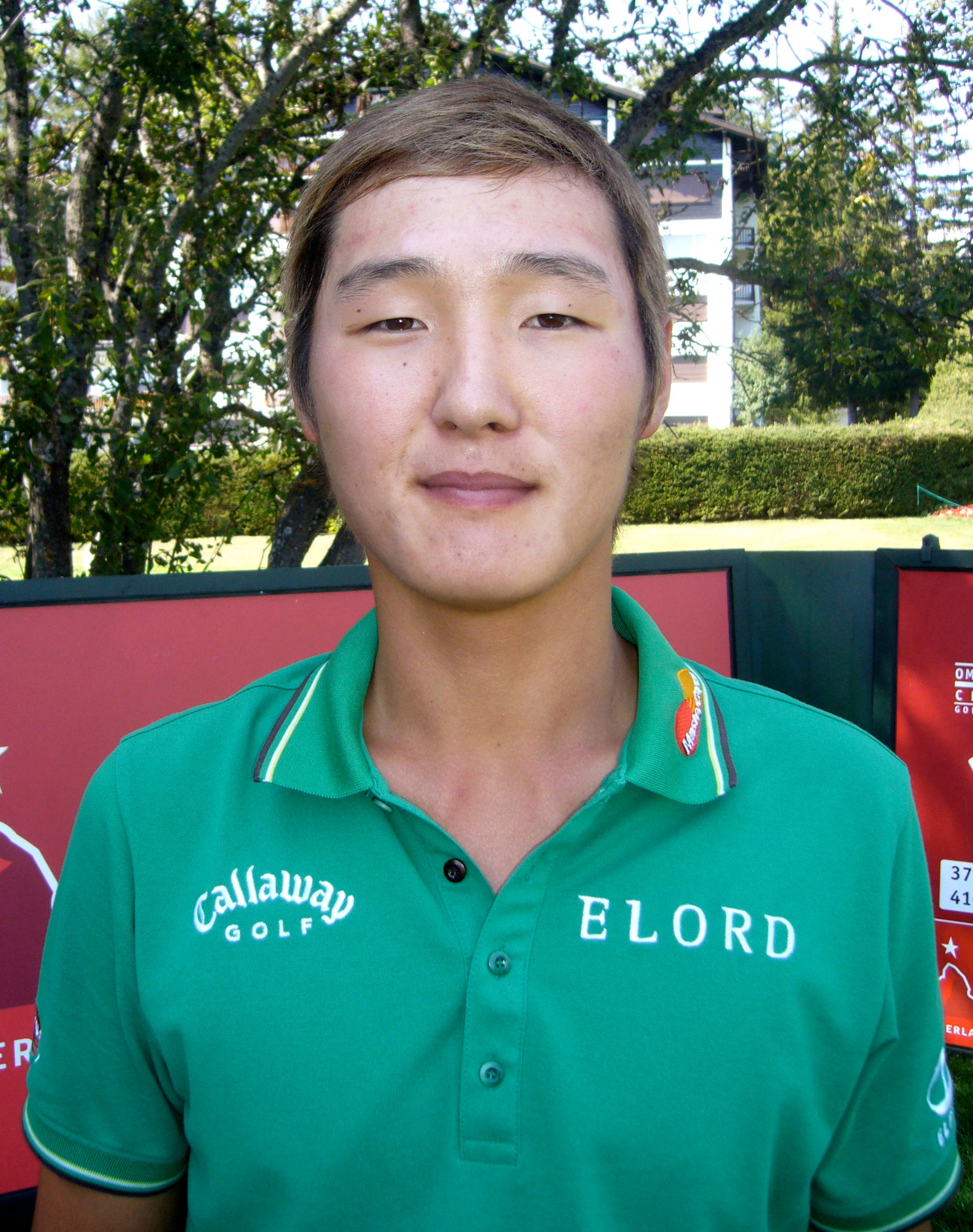 高爾夫 桌布 壁紙 — 一位年輕高爾夫球手穿著綠色Callaway Polo衫，戶外站立，身後有樹木和紅色橫幅。