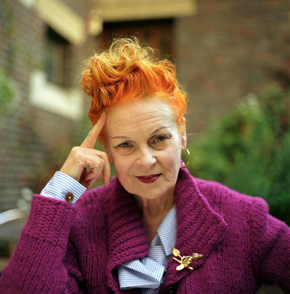 Vivienne Westwood 桌布 壁紙 — 一位年長女性，擁有火紅橙色頭髮，身穿紫色針織外套，在磚牆建築背景下若有所思地擺姿勢。
