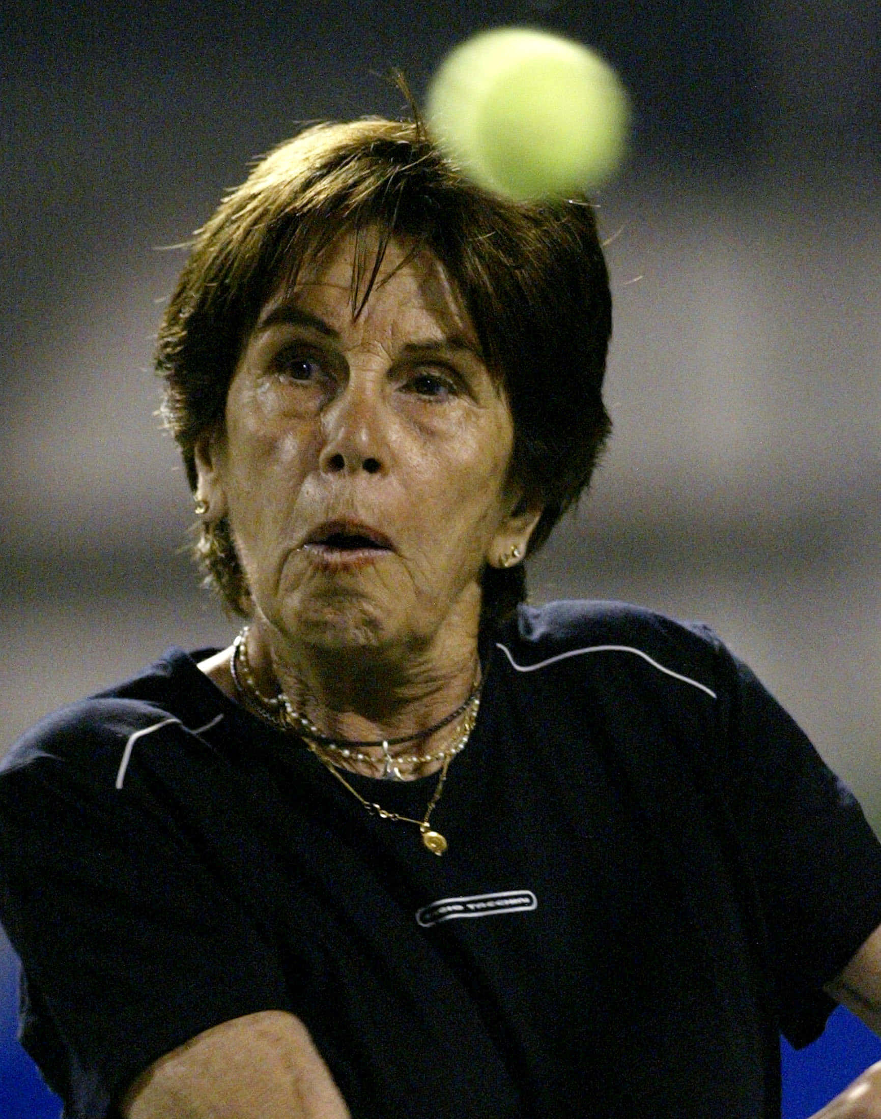 Tennis 桌布 壁紙 — 一位年長女性在夜間比賽中專注地揮動球拍，擊向模糊的網球。