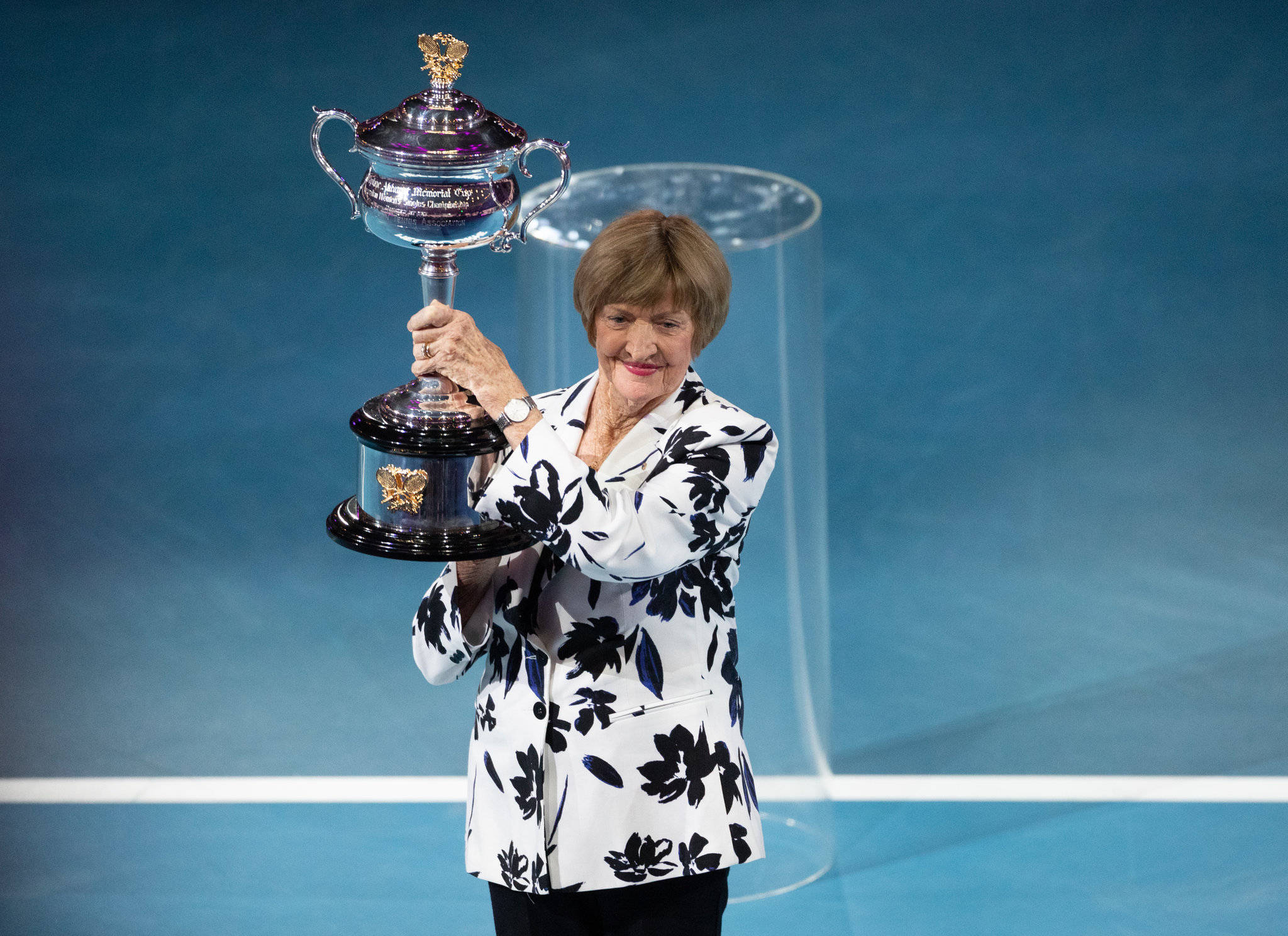 Australian Open 桌布 壁紙 — 一位年長女性穿著花卉圖案西裝外套，在藍色球場上自豪地舉起一個大型銀色網球獎盃。