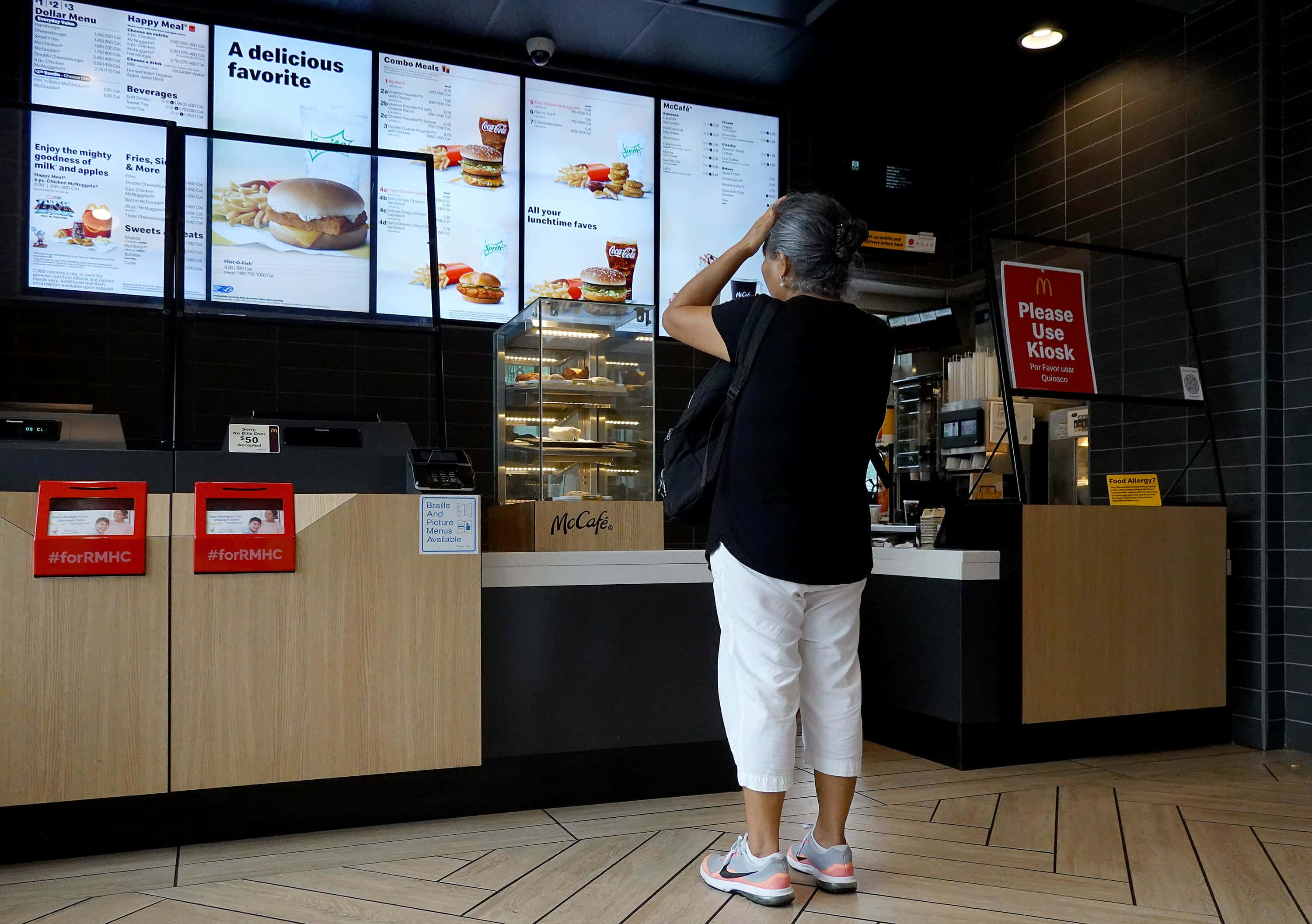 fast food 桌布 壁紙 — 一位年長女性站在麥當勞餐廳內，抬頭看著顯示食品項目的數位菜單看板。