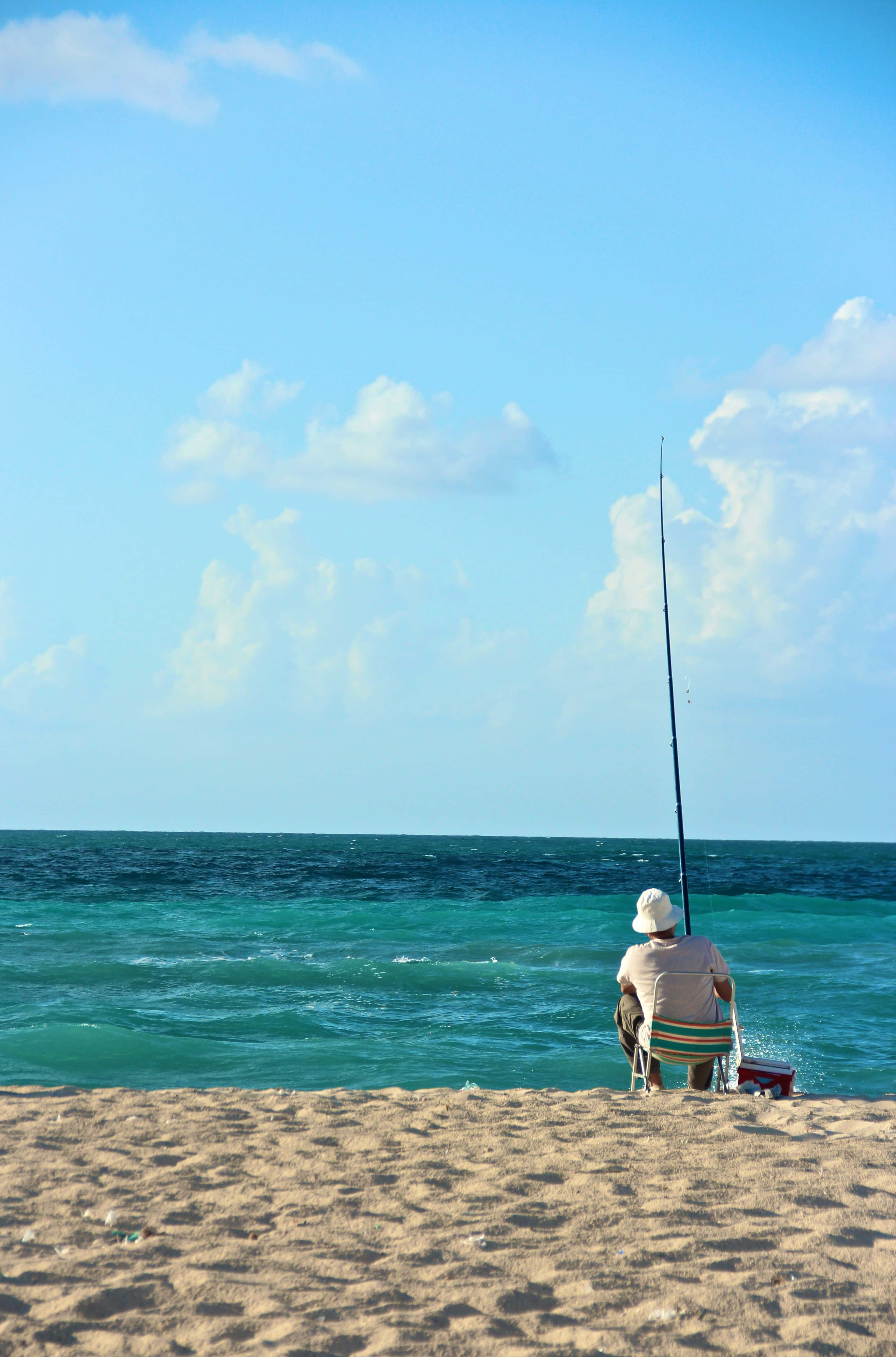 Fishing Phone 桌布 壁紙 — 一位年長漁夫獨自坐在沙灘上，在藍天白雲下將魚線拋入碧綠的海洋。