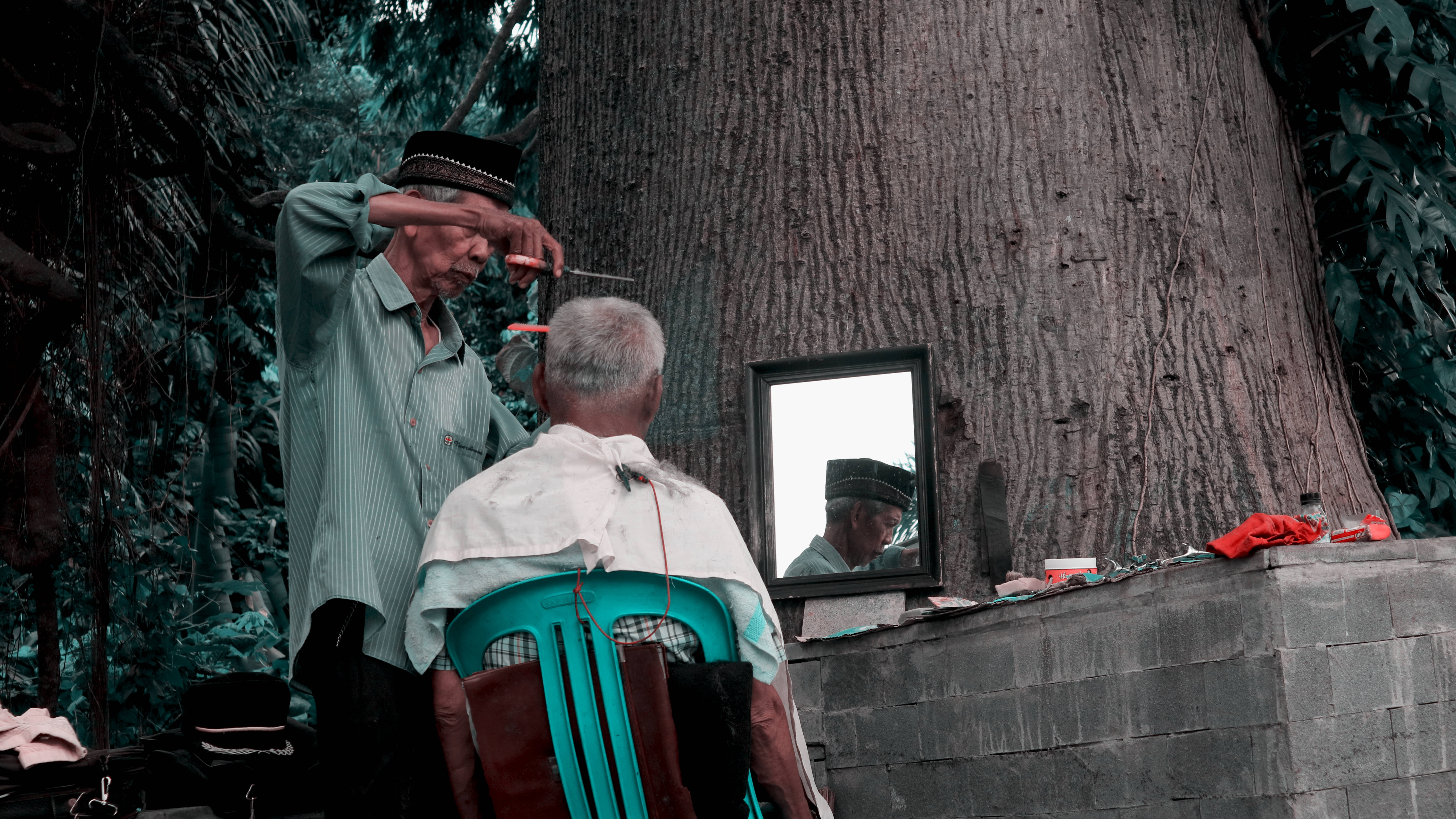 Haircut 桌布 壁紙 — 一位年長理髮師在戶外一棵大樹旁為客人修剪頭髮，鏡子映照出他專注的表情。