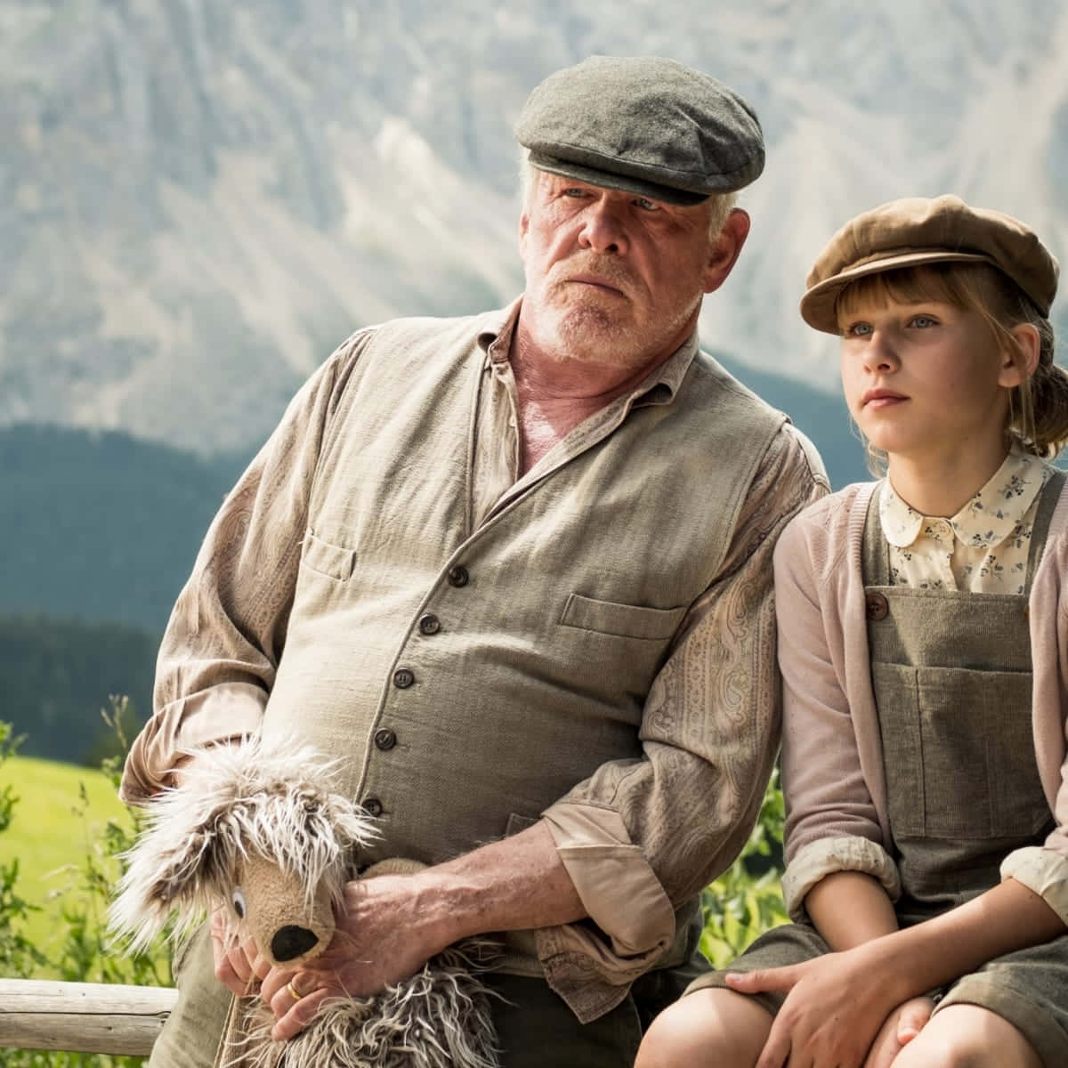 Nick Nolte 桌布 壁紙 — 一位年長男士和一位年輕女孩戴著復古帽子坐在一起，抱著一隻破舊的泰迪熊，背景是崎嶇的山脈和綠色的草地。