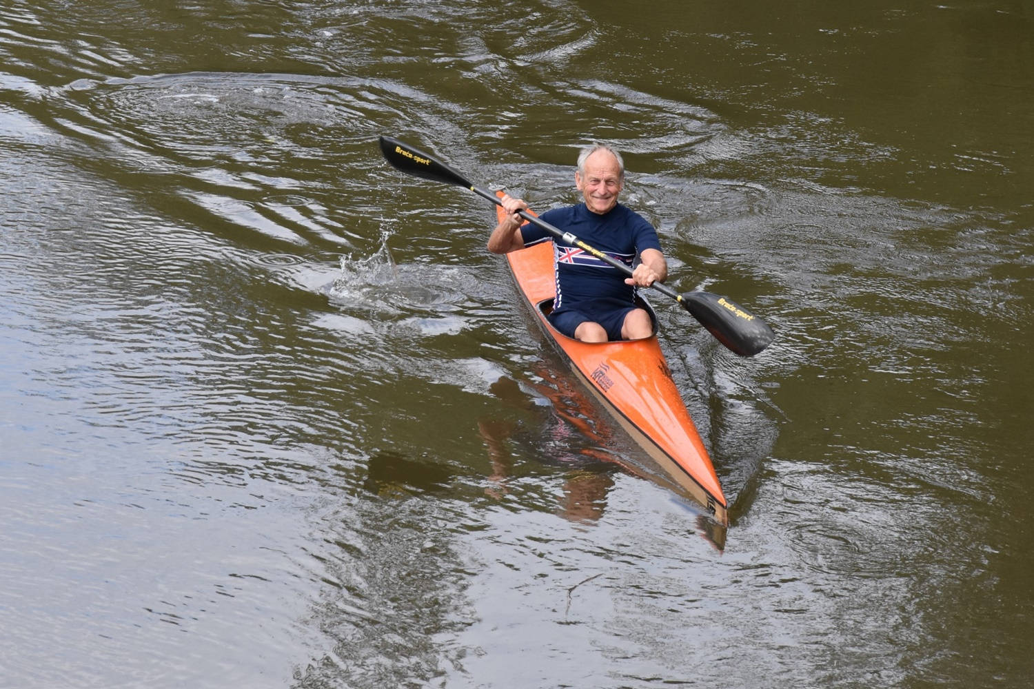 Rowing 桌布 壁紙 — 一位年長男士在平靜、泛起漣漪的河上划著橙色獨木舟，露出愉快的笑容。