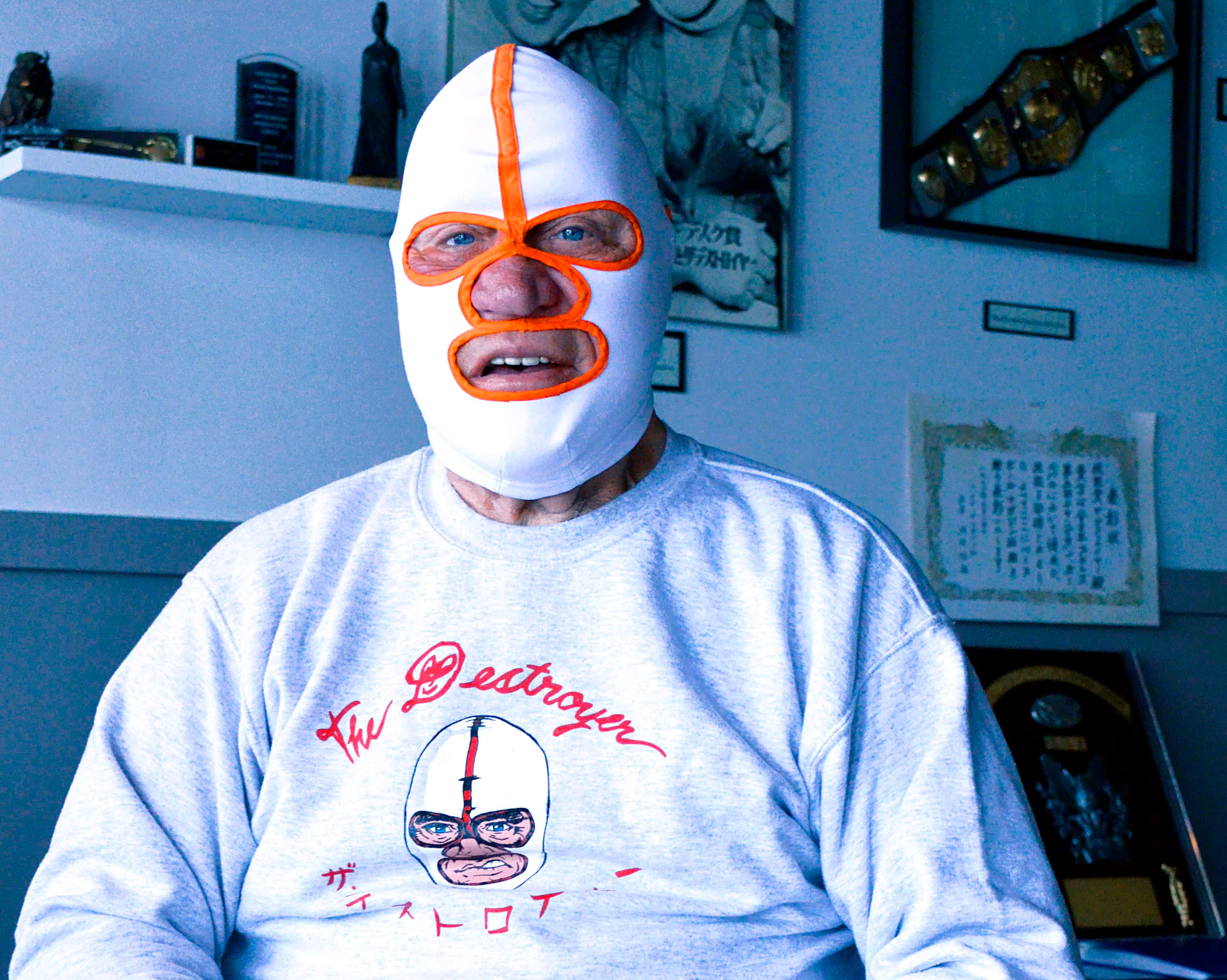 WWE 桌布 壁紙 — 一位年長男士戴著白色橙色邊的 lucha libre 面具，室內穿著印有自己面具形象的衛衣。