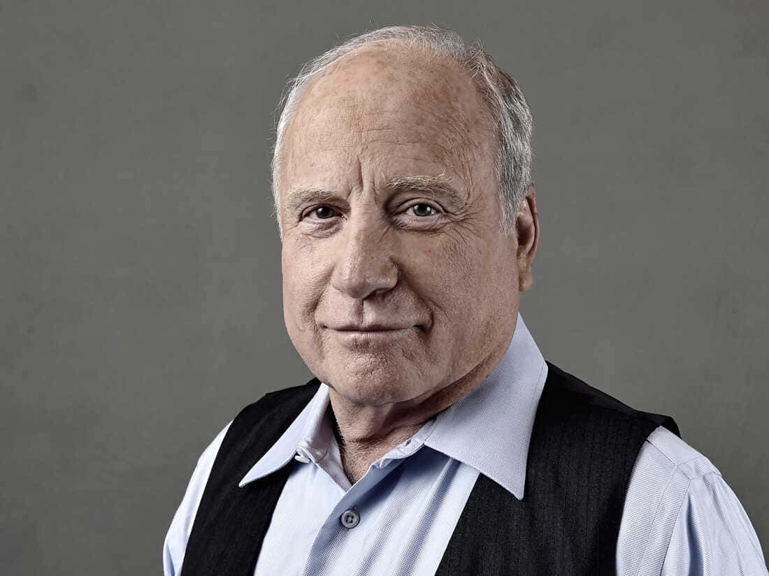 Richard Dreyfuss 桌布 壁紙 — 一位年長男士的close-up肖像，他帶著溫柔的微笑，穿著淺藍色襯衫和深色背心，背景為中性灰色。
