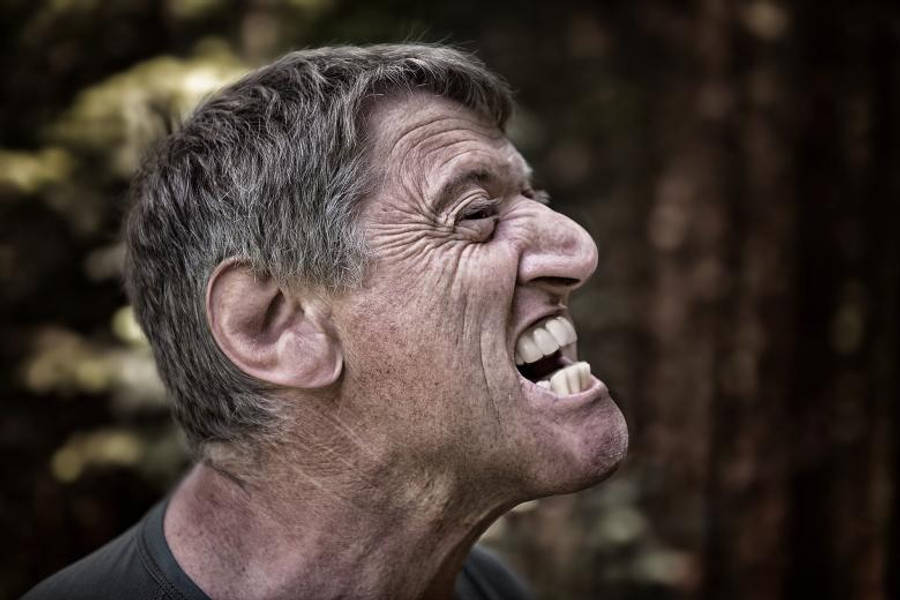 Angry 桌布 壁紙 — 一位年長男士的近距離特寫，他開懷大笑，臉上帶有深深的皺紋和灰白頭髮，背景是模糊的森林。