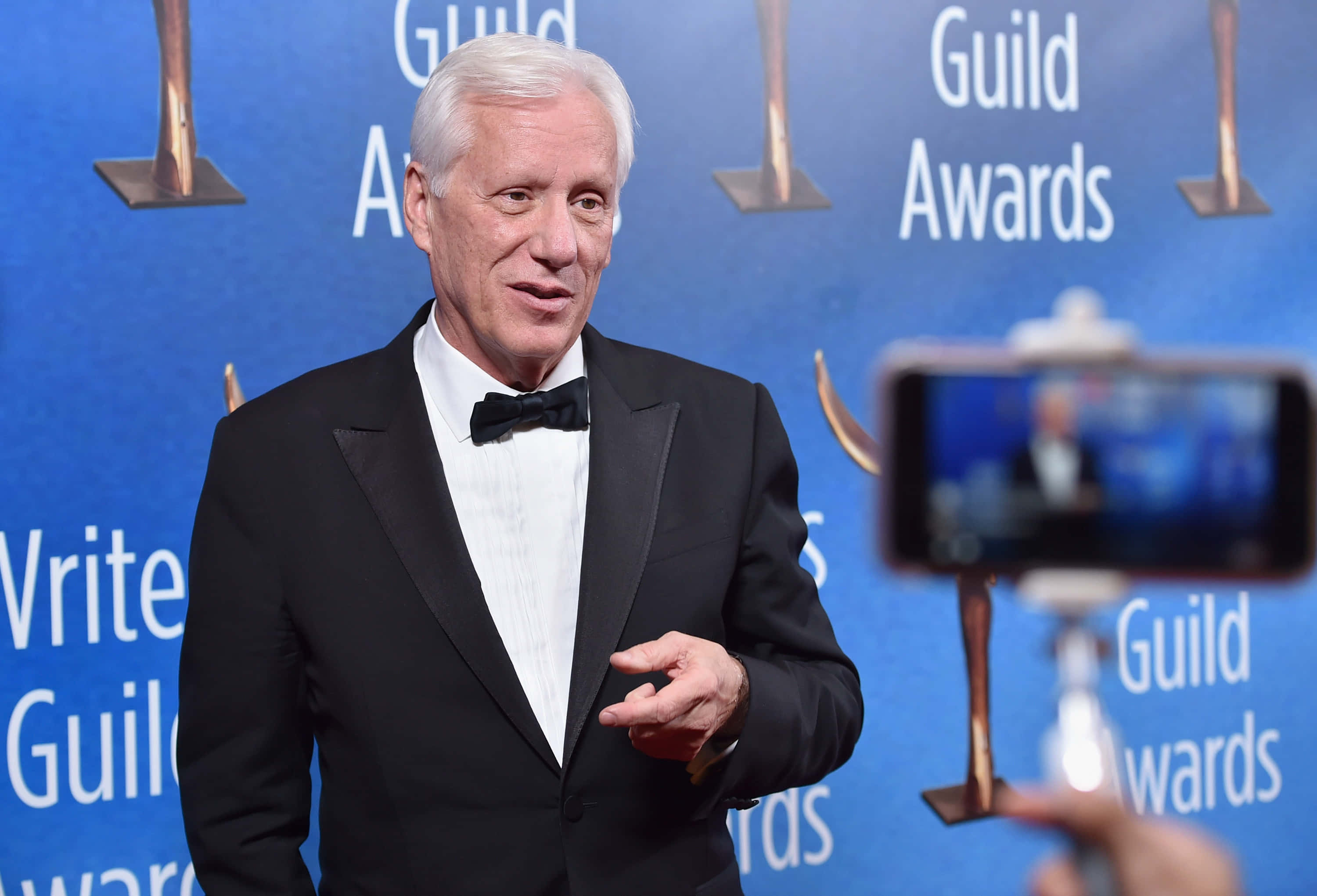 James Woods 桌布 壁紙 — 一位年長男士身穿黑色燕尾服和領結，在Guild Awards上發言，智慧型手機在藍色背景下錄製他。
