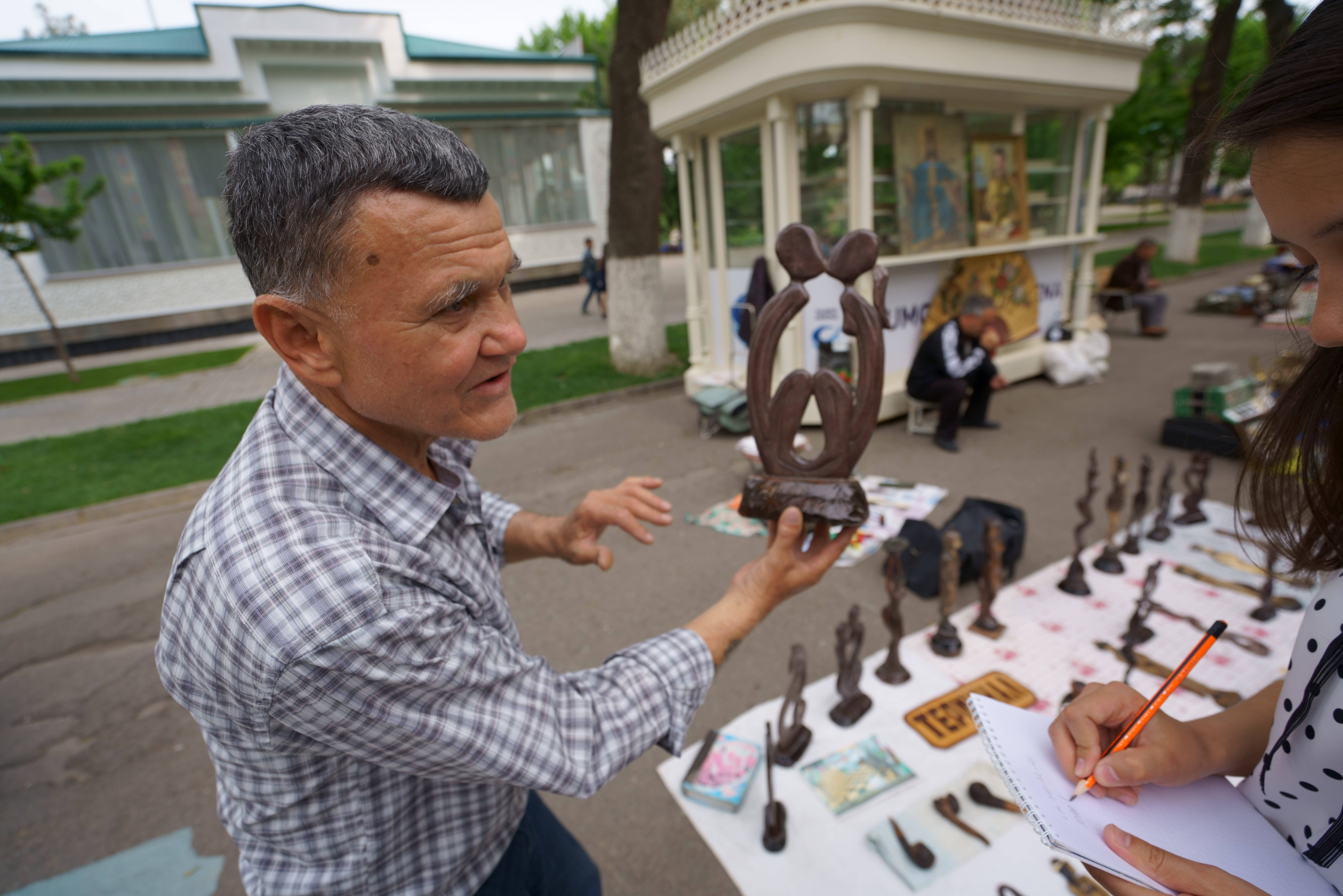 Uzbekistan 桌布 壁紙 — 一位年長男子在戶外工藝品市場熱情地向一位正在做筆記的年輕女子解釋一件青銅雕塑。