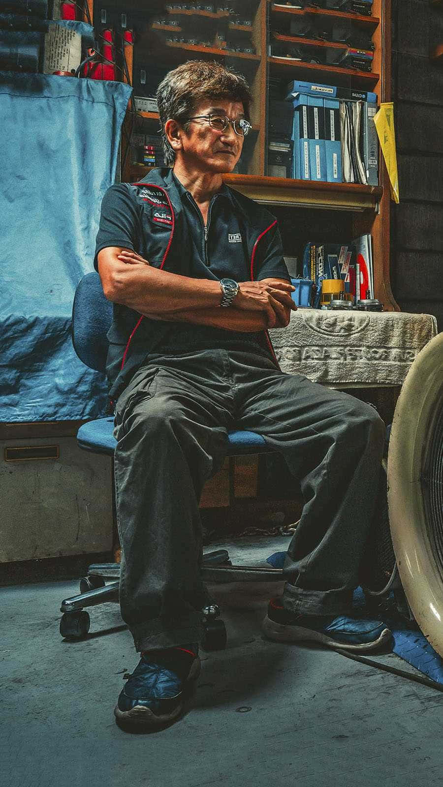 人像動態 桌布 壁紙 — 一位年長男子穿著賽車制服，自信地坐在雜亂的車庫中，身後有工具和架子。
