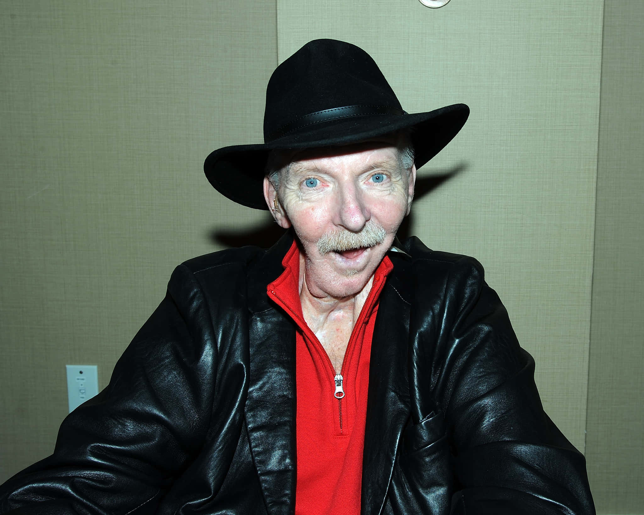 Bobby Heenan 桌布 壁紙 — 一位年長男性，擁有引人注目的藍色眼睛和鬍鬚，戴著黑色軟呢帽和皮夾克，內搭紅色襯衫，直視鏡頭。