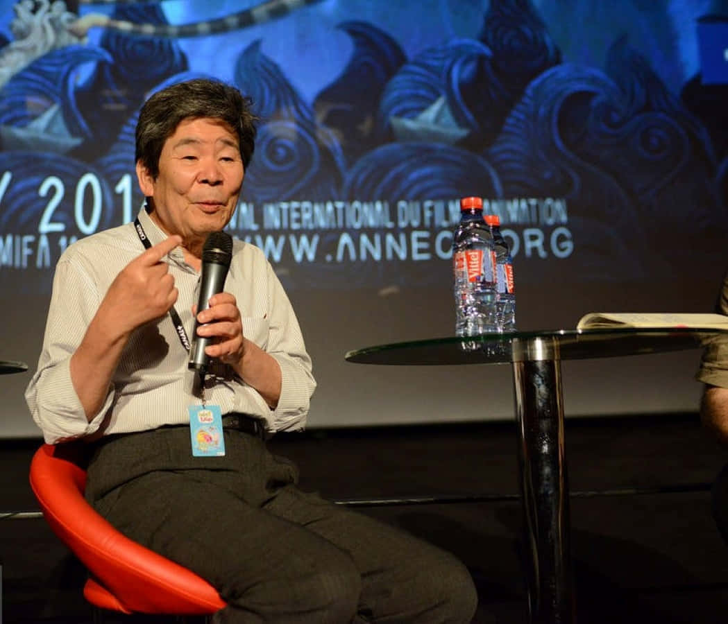 Isao Takahata 桌布 壁紙 — 一位年長男性坐在紅色椅子上，在動畫節座談會上對著麥克風發言，背景為藍色龍。
