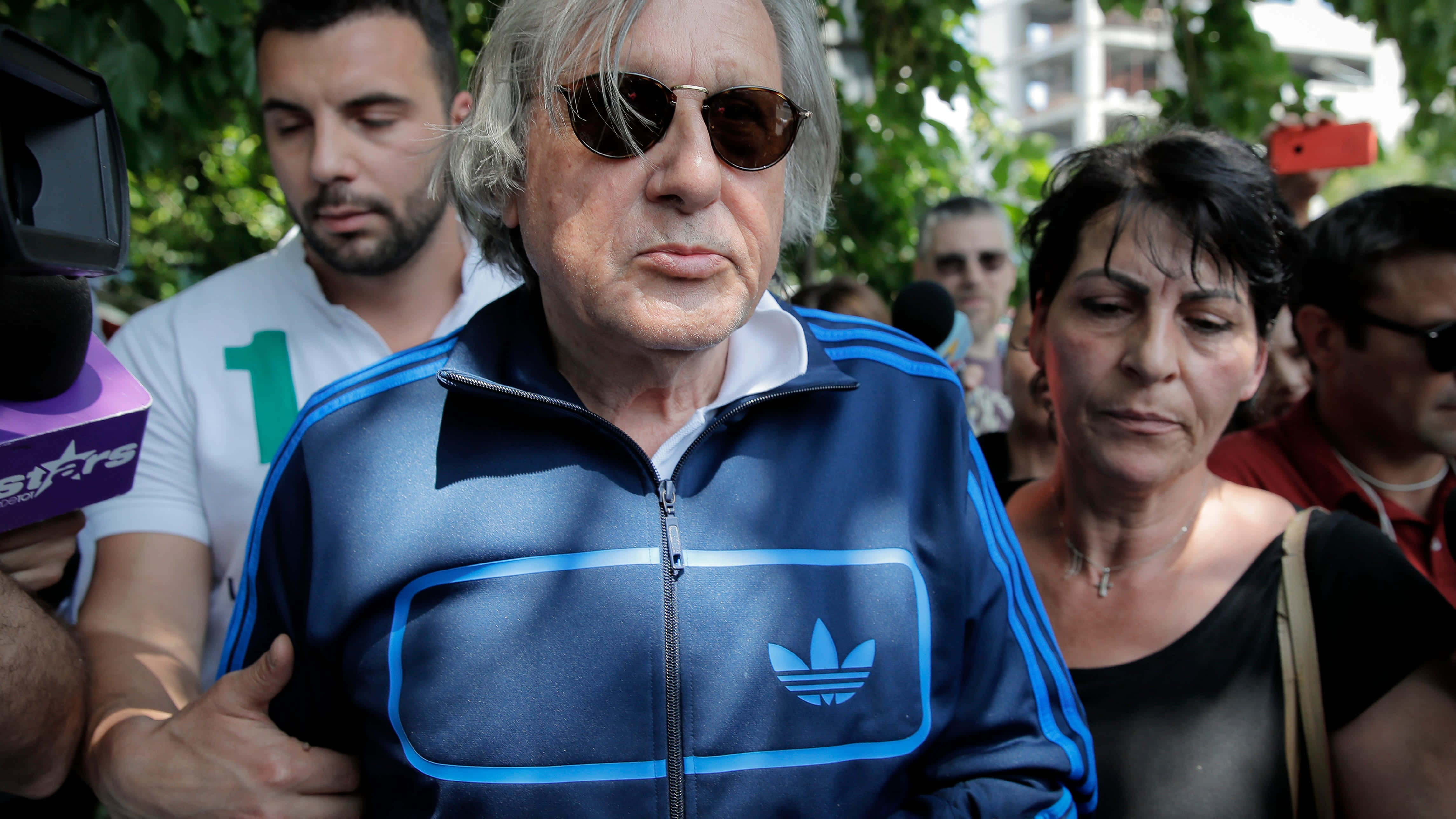 網球 桌布 壁紙 — 一位年長男性穿著藍色Adidas運動服和太陽眼鏡，在戶外穿過一群記者和圍觀者。