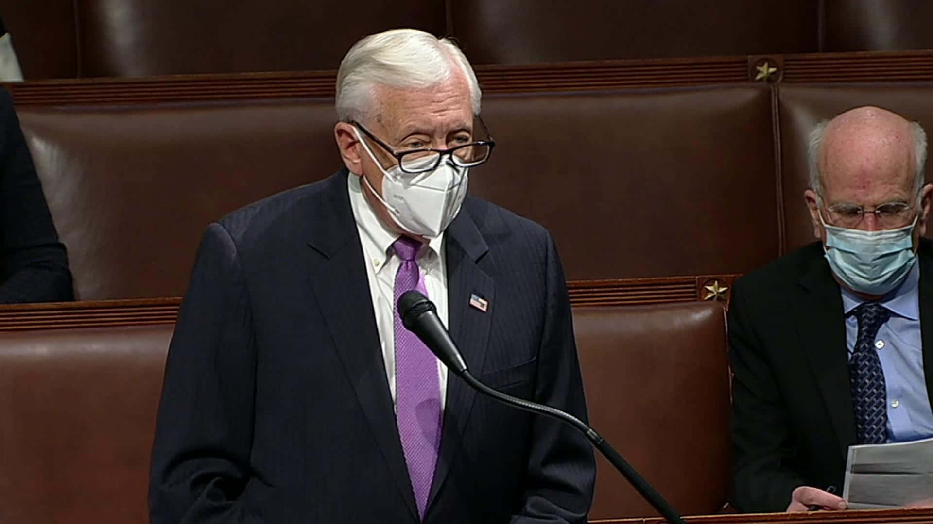 政治 桌布 壁紙 — 一位年長男性身穿深色西裝和紫色領帶，在正式的立法會議廳內戴著白色口罩對著麥克風發言。