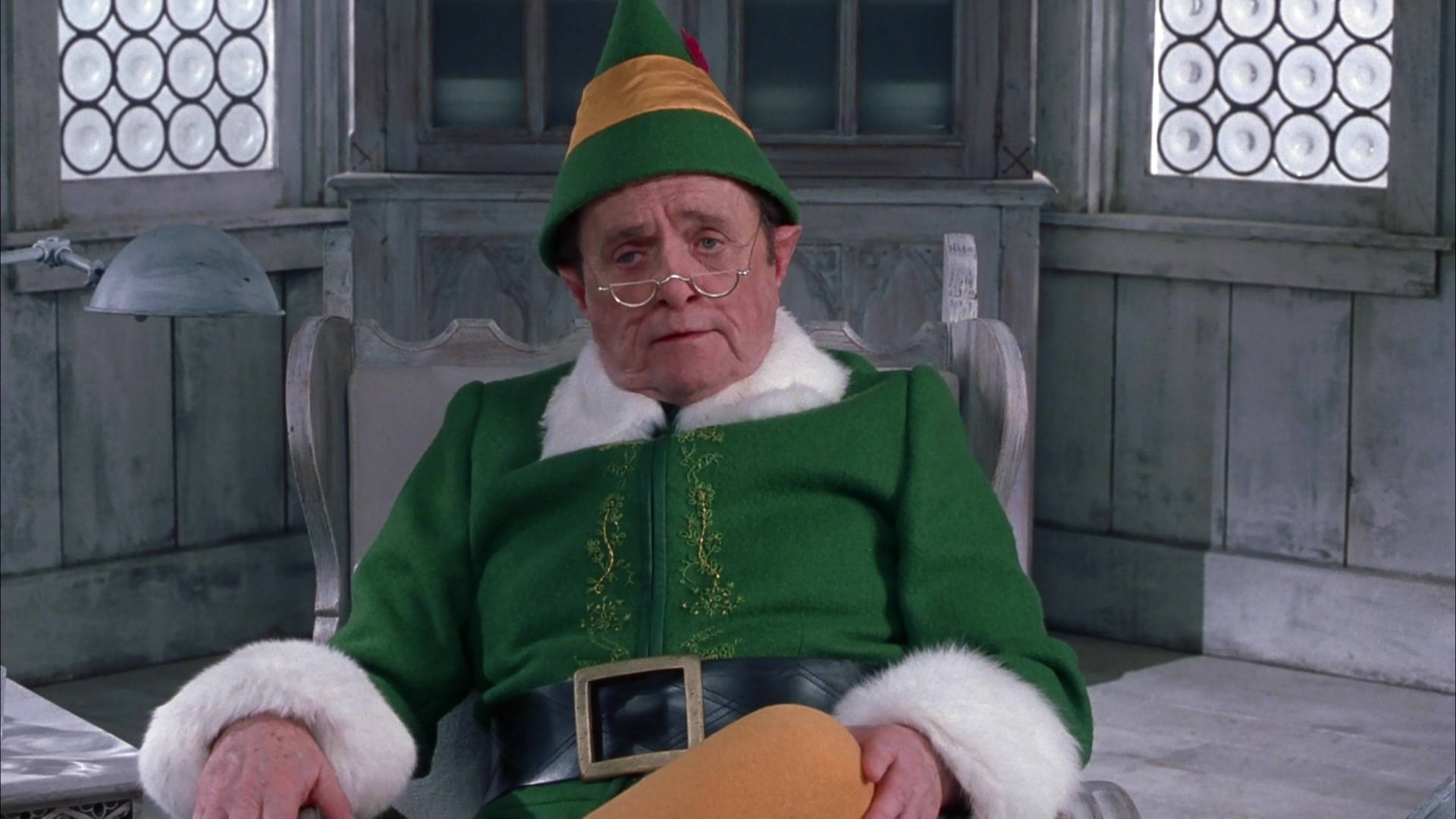 Elf Movie 桌布 壁紙 — 一位年長男性身穿綠色精靈服飾，飾有白色毛邊和尖頂帽，坐在鄉村小屋內的木椅上。