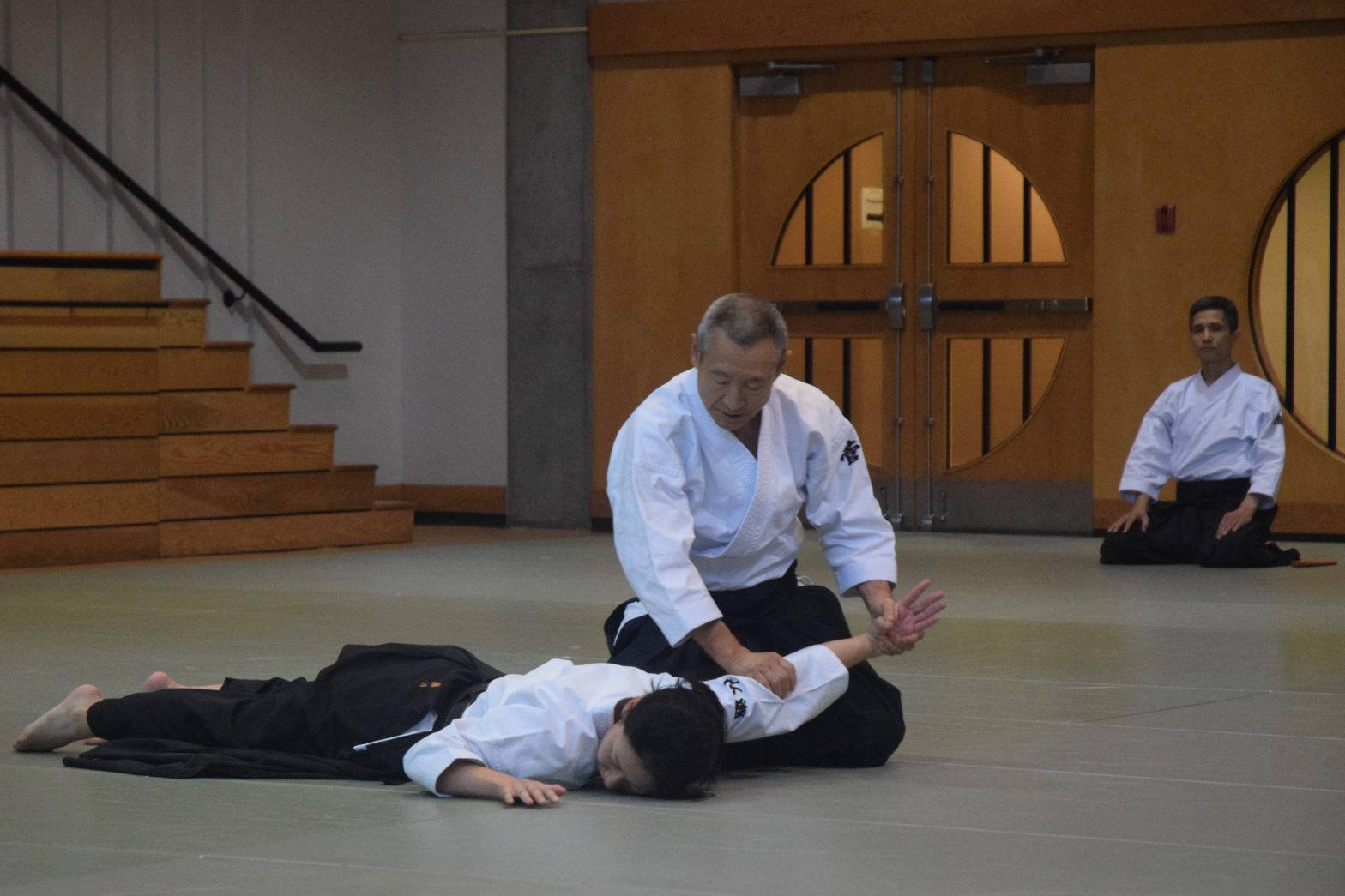martial arts 桌布 壁紙 — 一位年長的武術教練在一個有木門和樓梯的道場中，向學生演示纏鬥技巧。
