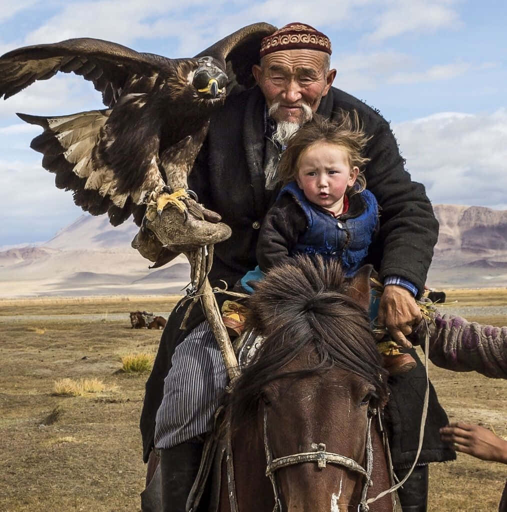 Mongolian 桌布 壁紙 — 一位年長的獵鷹人，膝上坐着一個孩子，騎着馬穿過廣闊乾旱的草原，天空多雲，一隻雄偉的金雕棲息在他戴着手套的手上。
