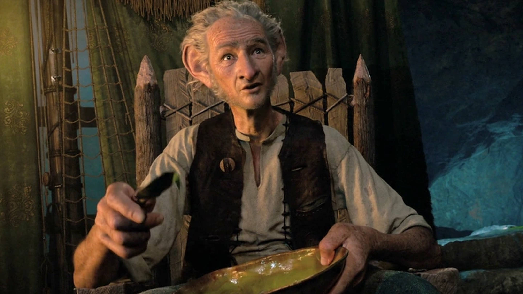 Bfg 桌布 壁紙 — 一位年長的精靈，尖耳朵，坐在木椅上，拿著碗和湯匙，在光線昏暗的小屋裡，有綠色窗簾和一個顯示暴風雨海洋的窗戶。